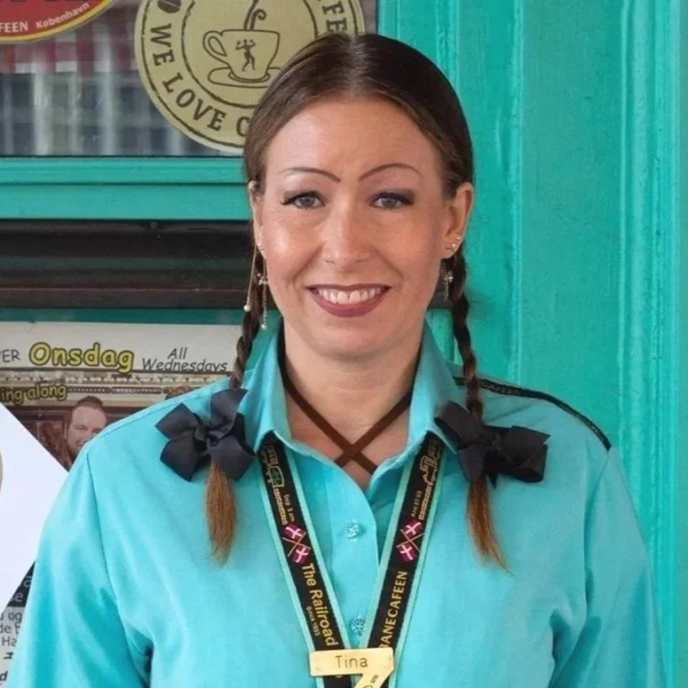 En smilende kvinde med to fletninger iført blå uniform og navy bøjler, med navn 'Tina' på en gul navneplade. Baggrunden er turquoise.