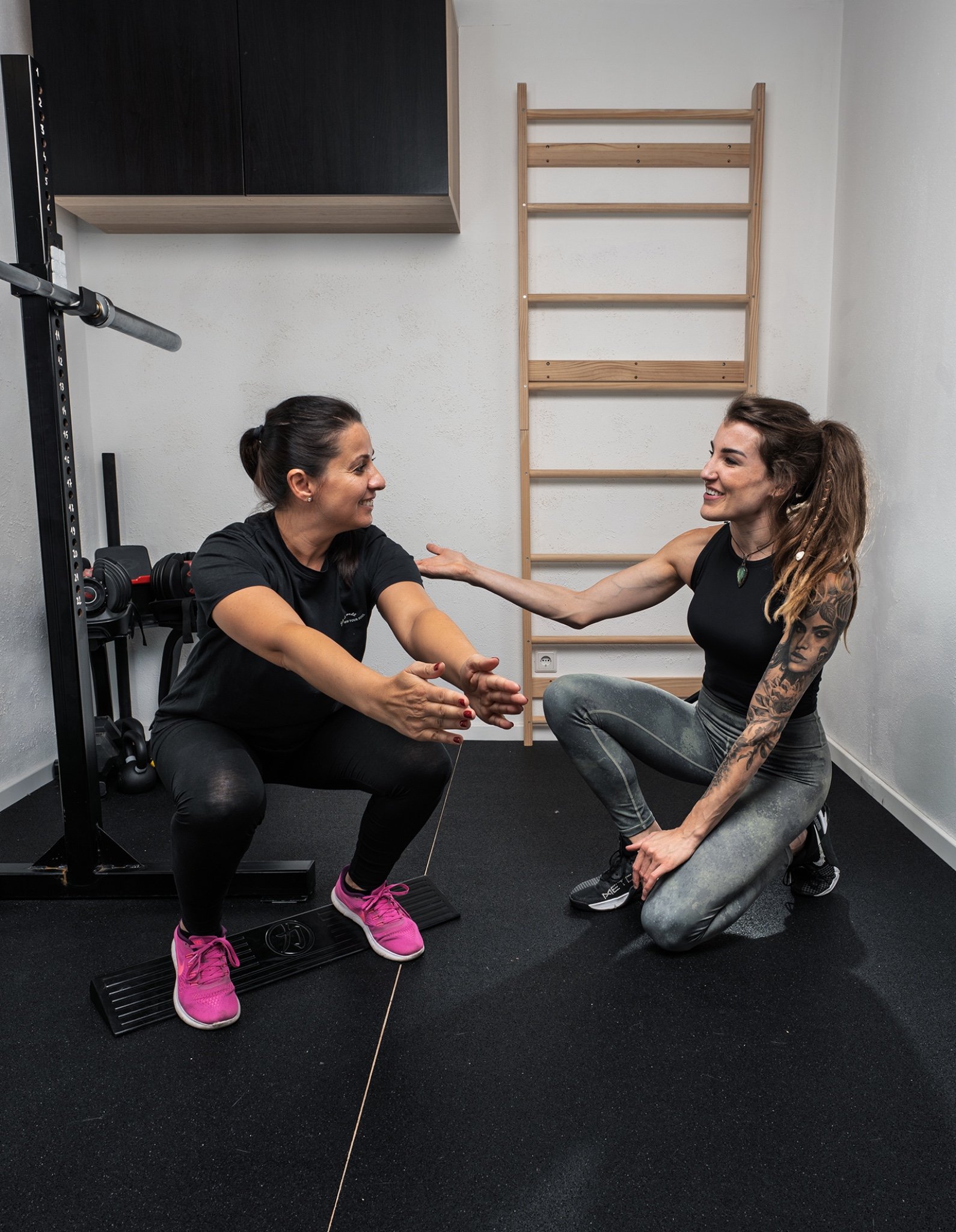 Zwei Frauen trainieren im Fitnessstudio, eine macht Kniebeugen, die andere gibt Anweisungen.