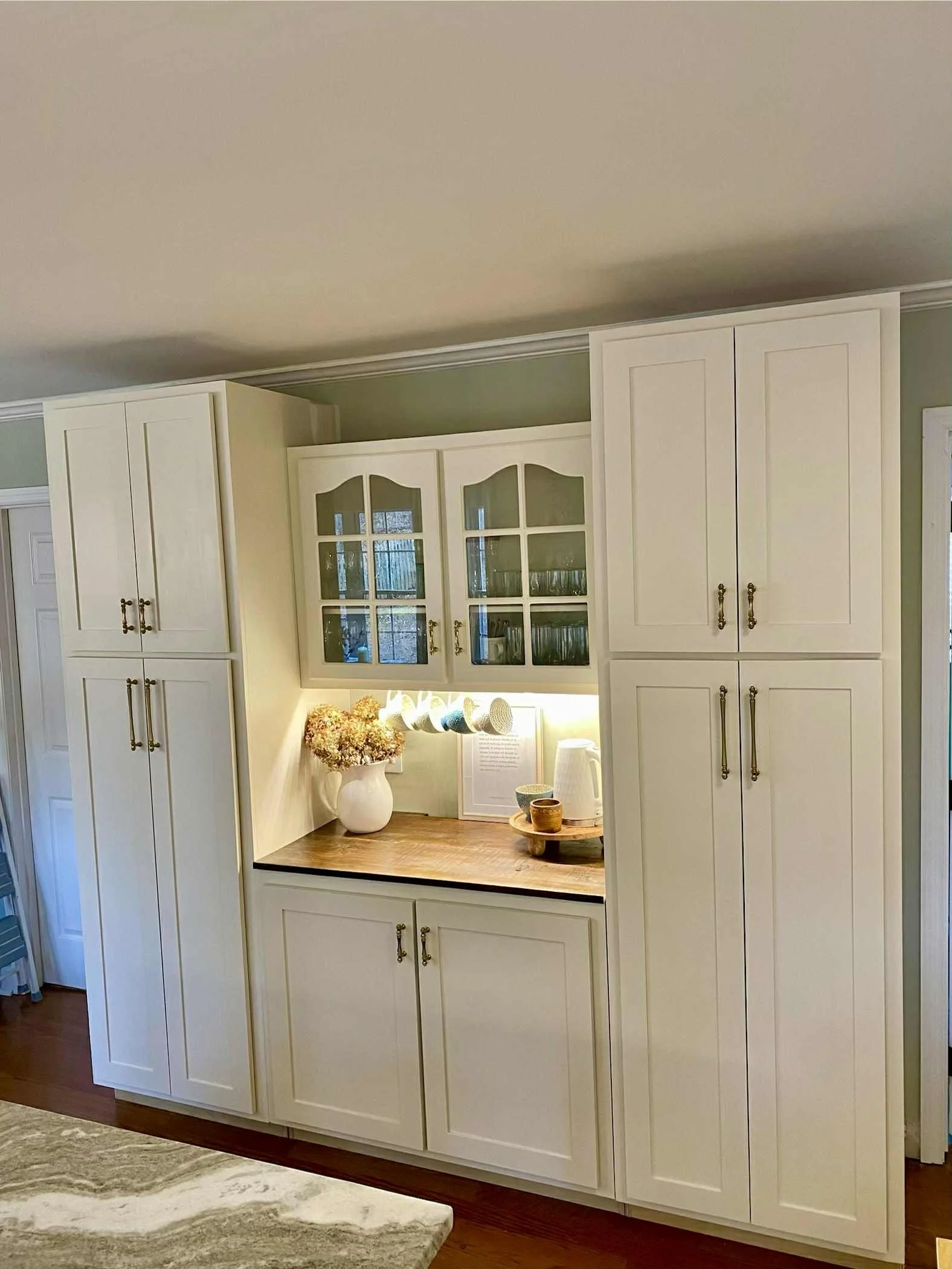 White kitchen cabinets with glass-front upper doors, a wooden countertop, a vase of flowers, a framed text, a white pitcher, and cups.
