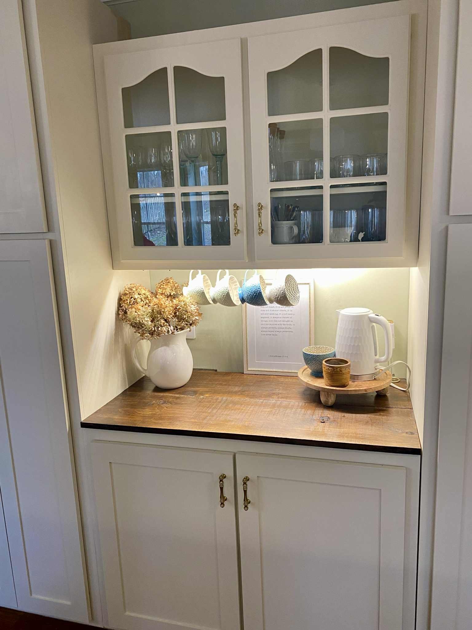 A kitchen cabinet with glass doors above a wooden countertop, decorated with a white vase of dried flowers, cups hanging below, and a tray with cups and a kettle. A framed paper stands behind the tray.