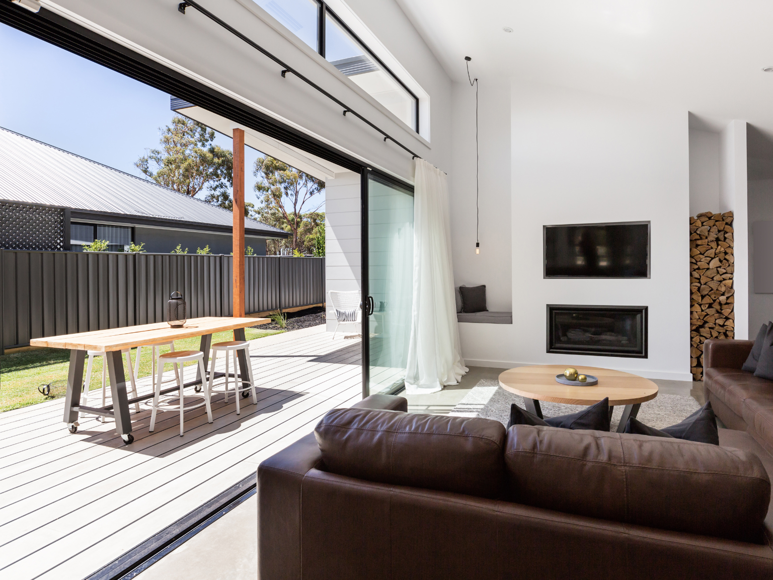 Living room with a brown leather sofa, round wooden coffee table, wall-mounted TV, fireplace, stacked firewood, built-in window seat with cushions, and large glass sliding door opening to a deck with outdoor furniture and a backyard.