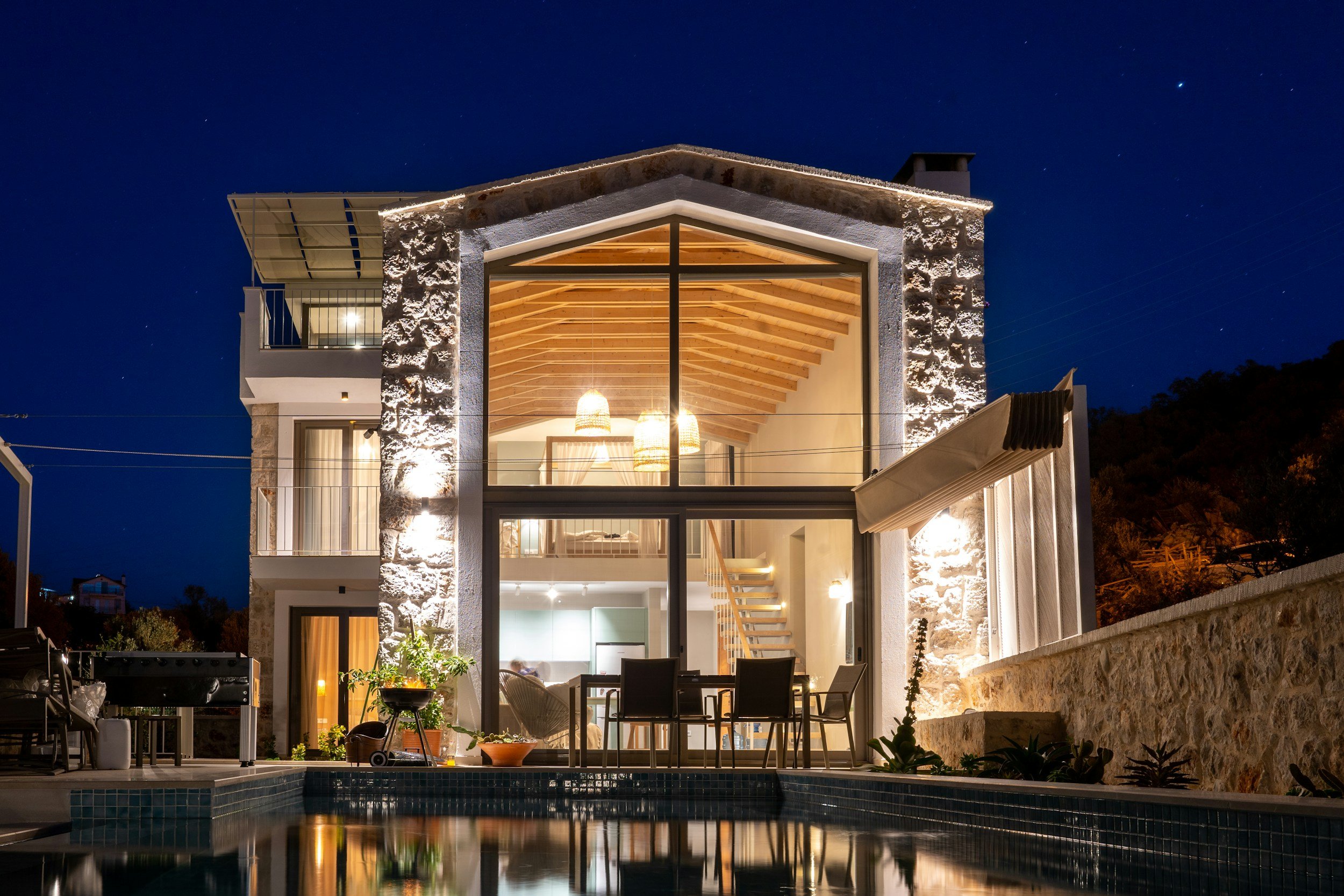 Night view of a modern, two-story house with large glass windows revealing a well-lit interior, stone and white exterior walls, and a swimming pool reflecting the house. The house has a sloped wooden roof, balcony, and outdoor seating area.