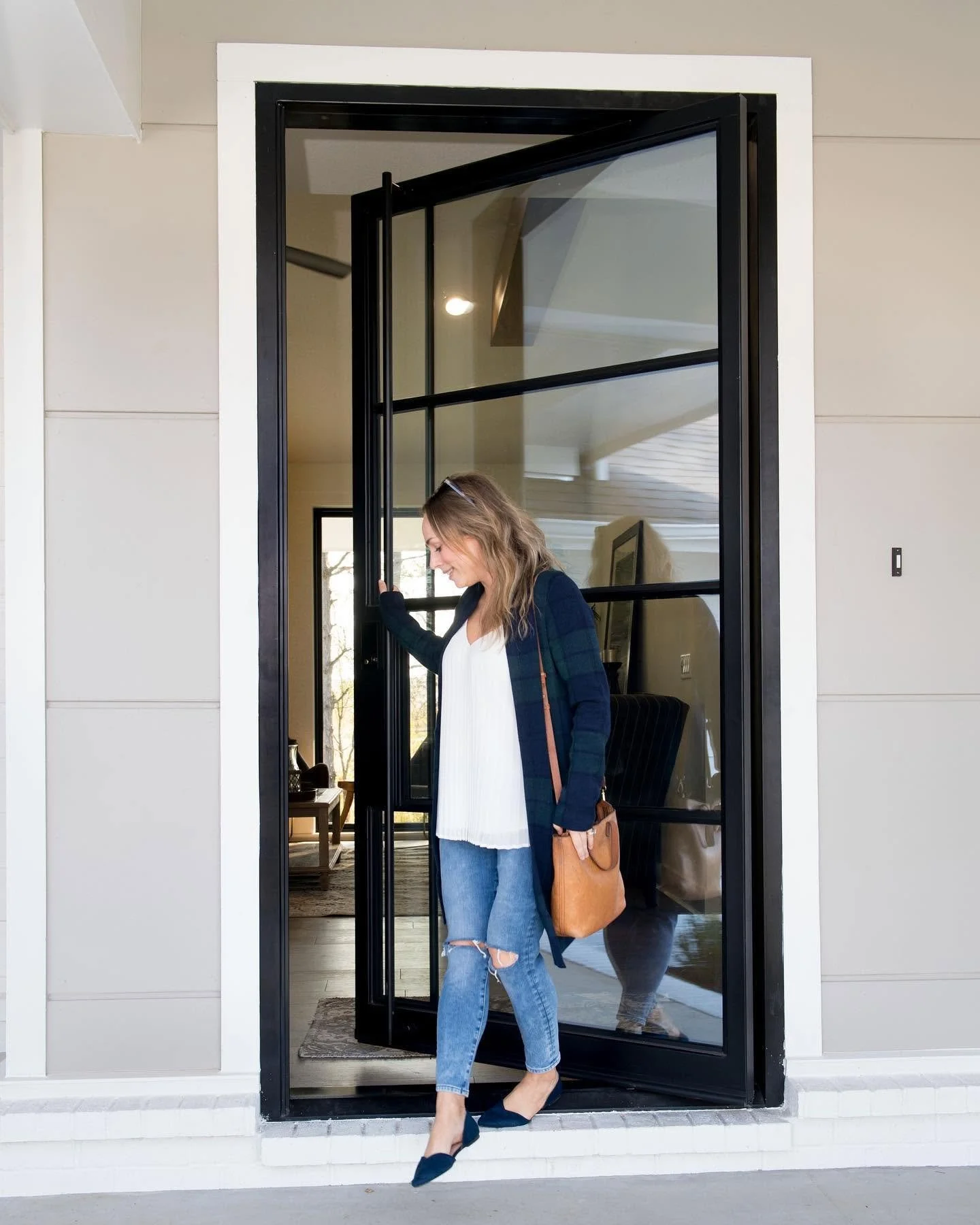 A woman with wavy blonde hair, wearing a white blouse, ripped jeans, and a dark plaid cardigan, steps out of a modern house through a black-framed glass door. She carries a tan tote bag and wears black pointed flats.