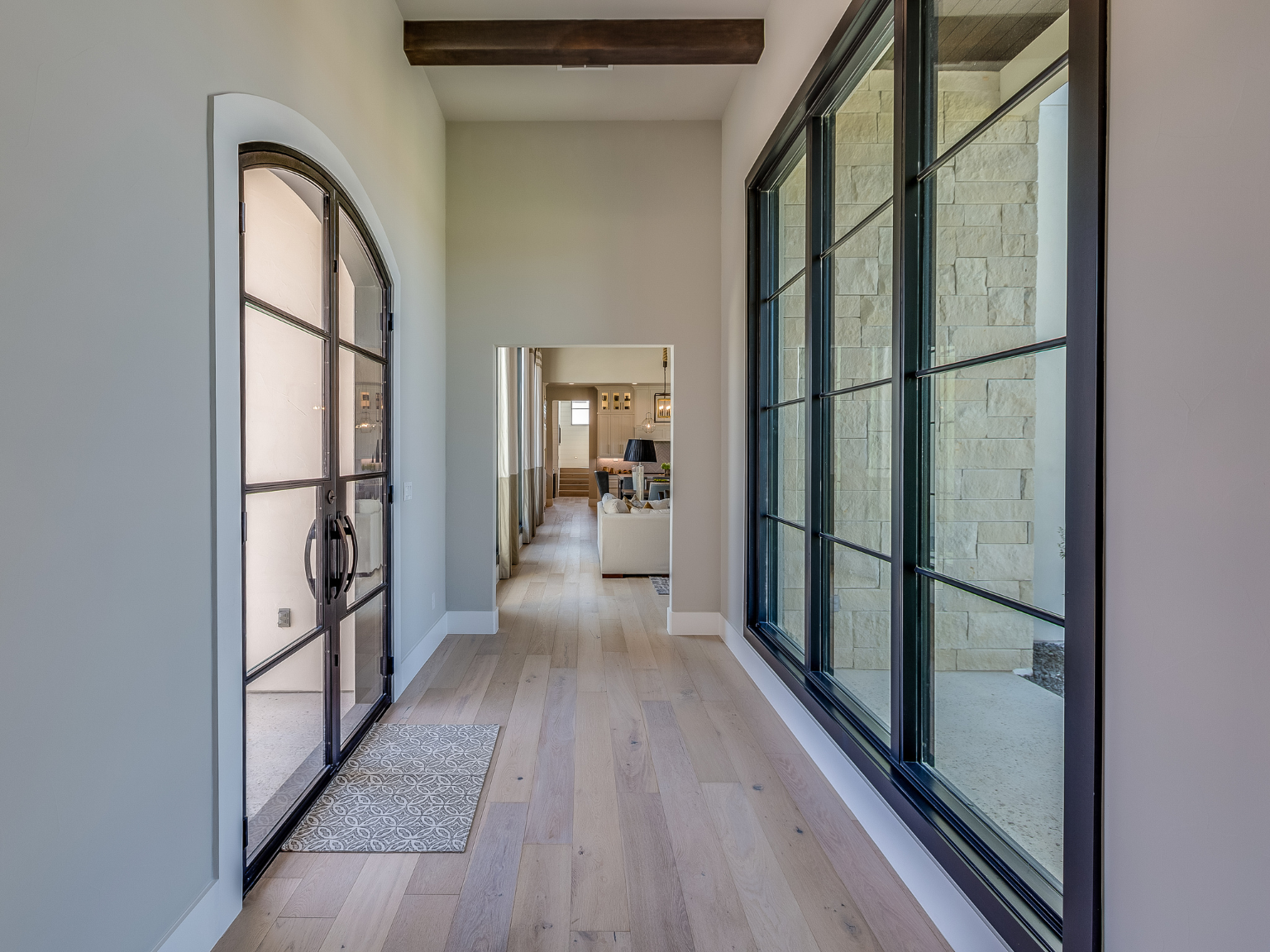 Interior view of a modern home entrance hallway with large window on the right, arched glass door on the left, and a view into the living room ahead, featuring light-colored hardwood floor, white walls, and a wooden ceiling beam.