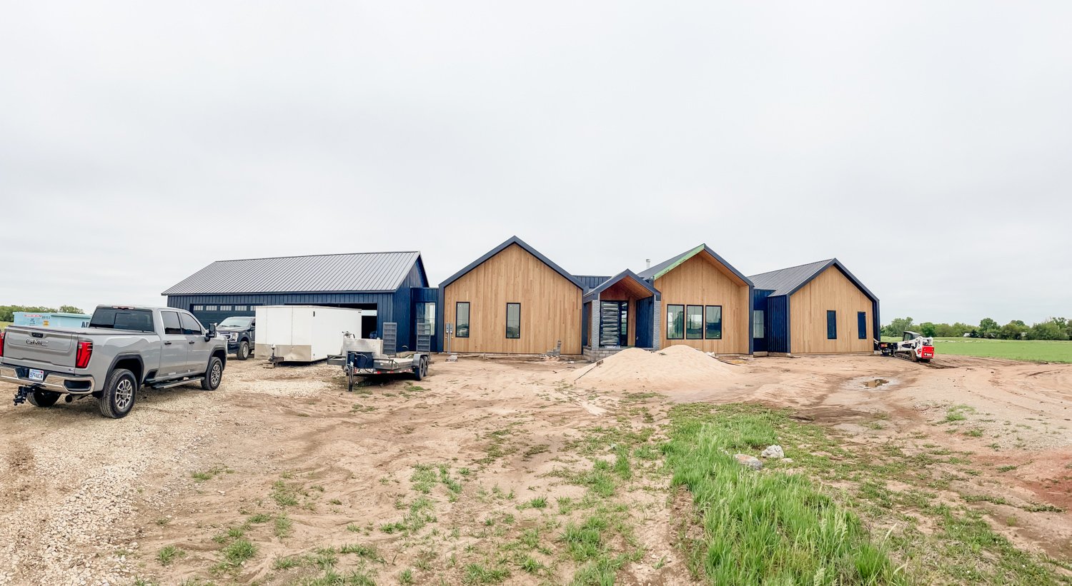 Under construction house with wooden and blue siding, construction vehicles parked nearby, dirt for landscaping, and an open field in the background.