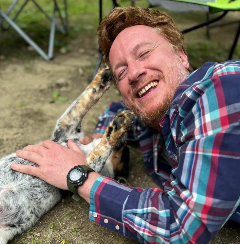 A man with red hair and a beard smiling and lying on the ground, playing with a dog outdoors. The man is wearing a colorful plaid shirt and a black wristwatch. The dog is lying on its back with paws in the air.