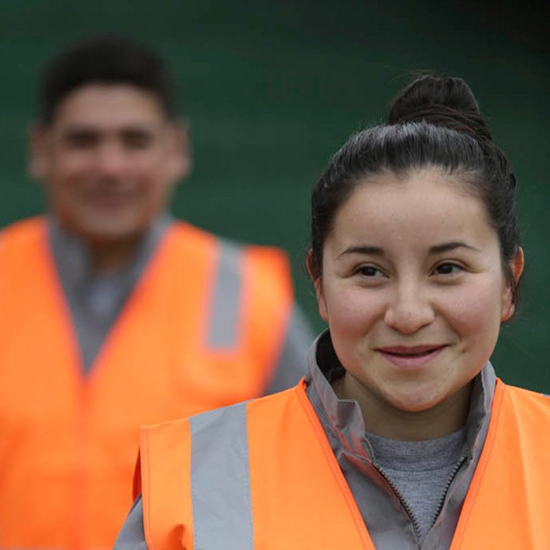 Mulher jovem sorrindo, usando colete de segurança laranja, com homem ao fundo também de colete, em ambiente ao ar livre.