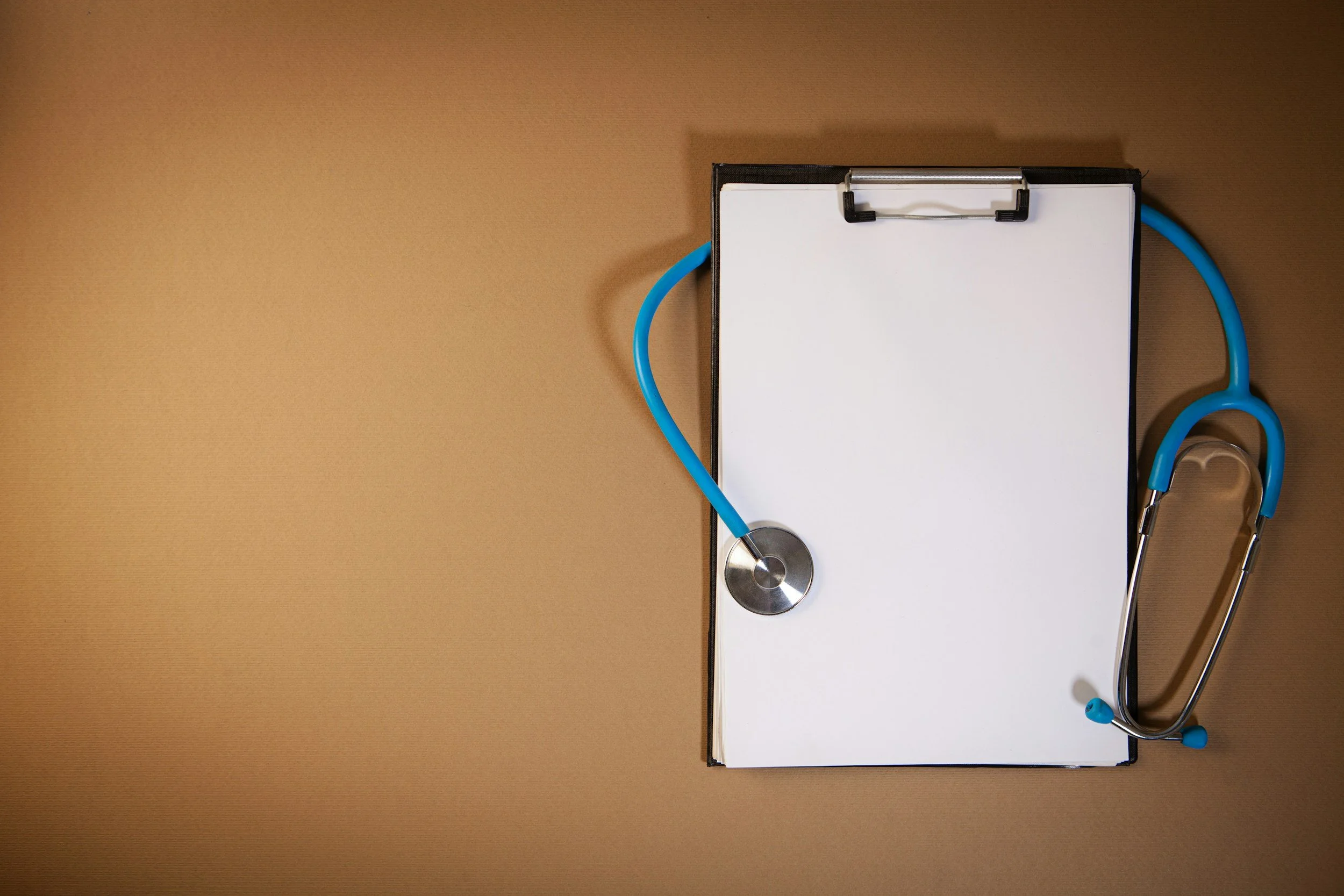 A medical clipboard with a blank sheet of paper, a blue stethoscope, and a silver stethoscope head placed on a brown surface.