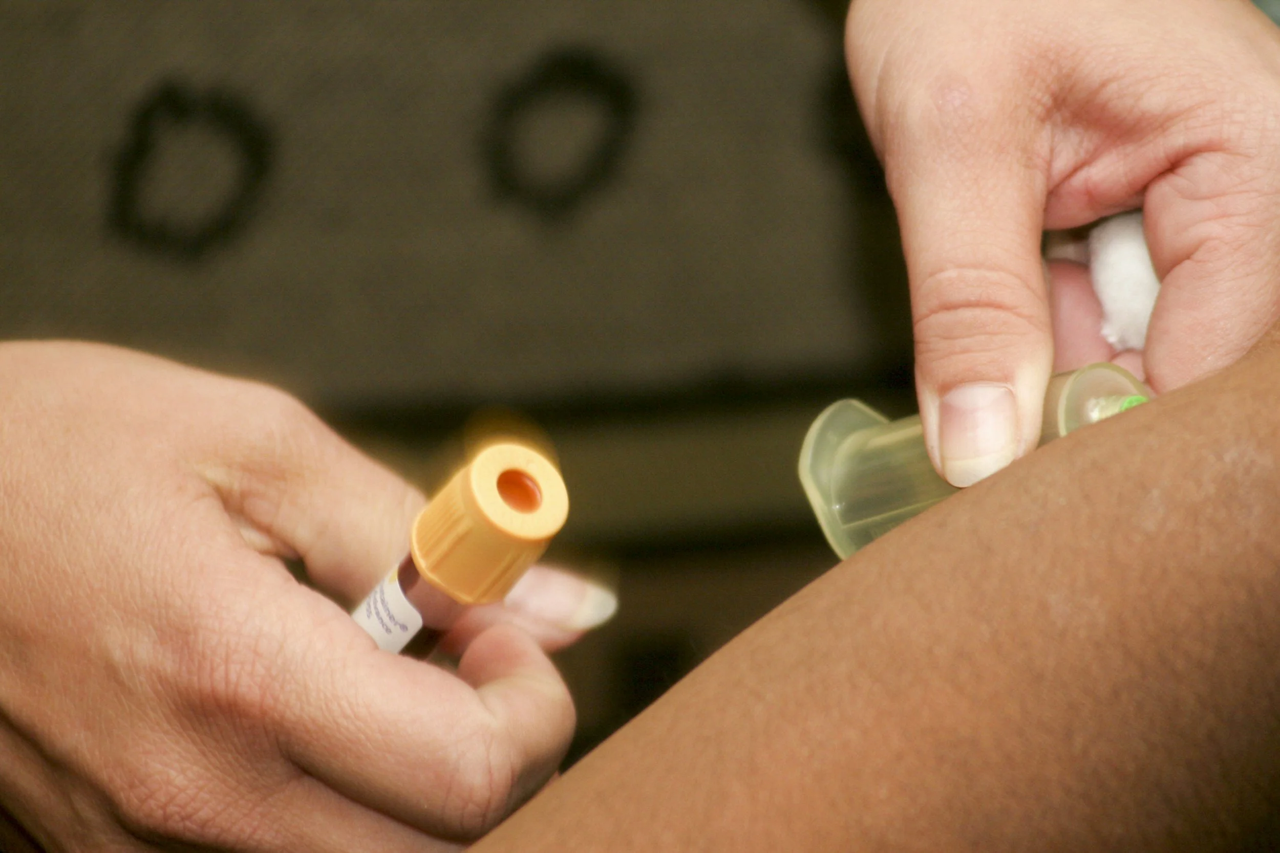 A close-up of a person administering an injection into another person's arm, holding a syringe and a cotton ball for lab draw