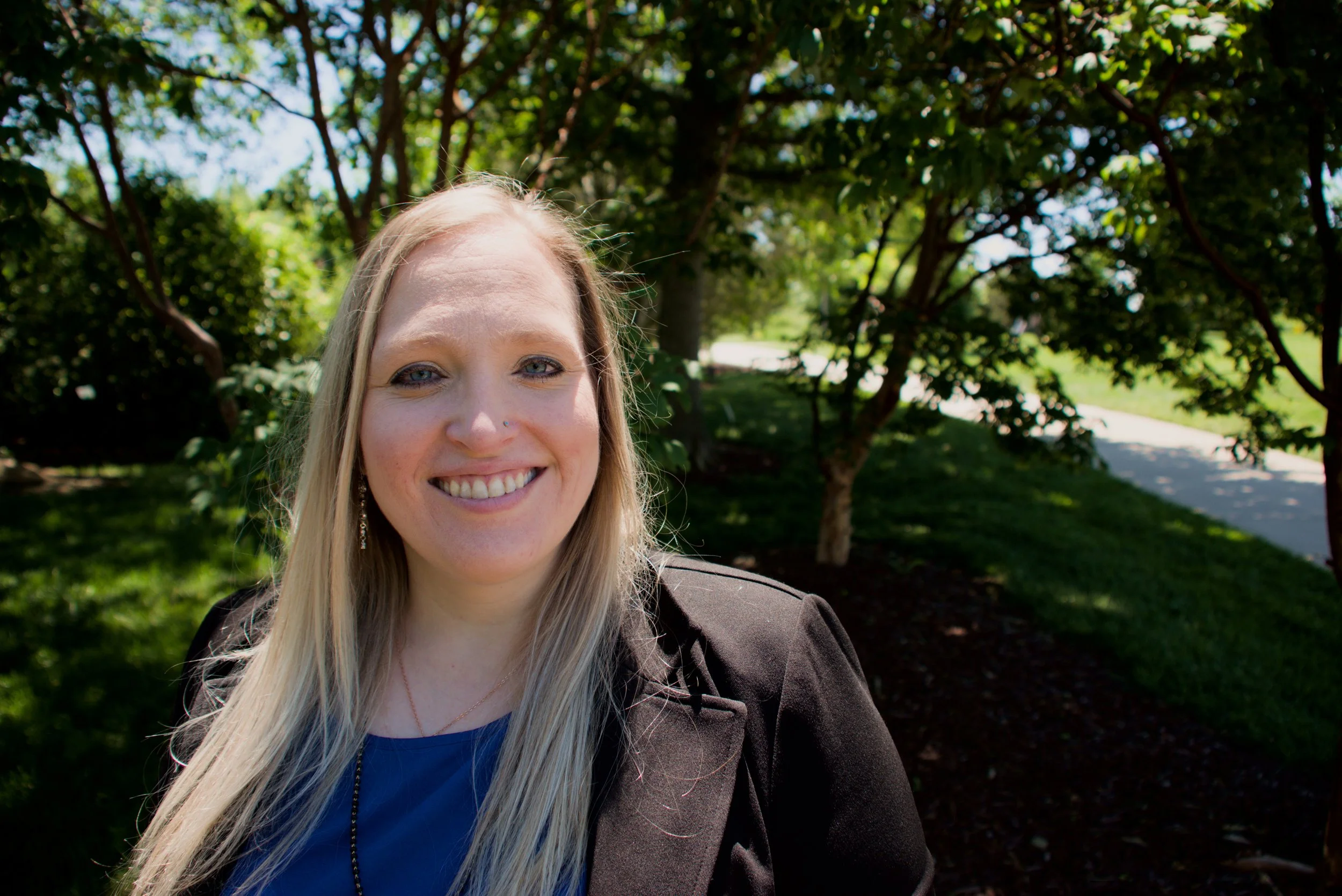 A smiling woman with long blonde hair taking a selfie outdoors in front of green trees and a pathway on a sunny day.