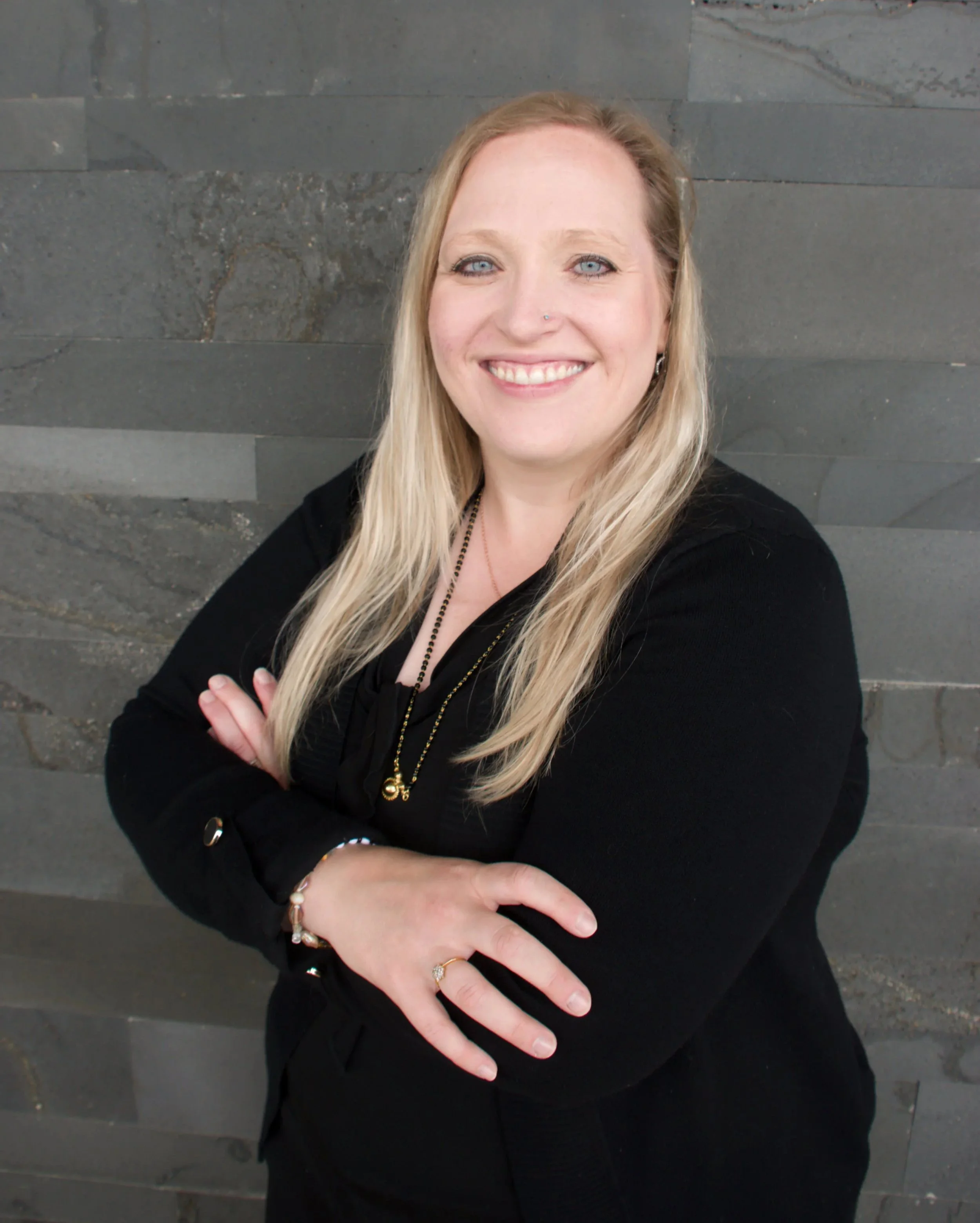 A woman with long blonde hair and blue eyes smiling with her arms crossed, standing against a gray stone wall.
