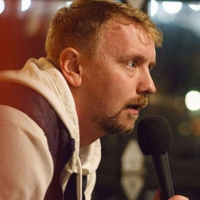 A man holding a microphone, appearing to speak at an event; he is wearing a white hoodie and looking intently forward.