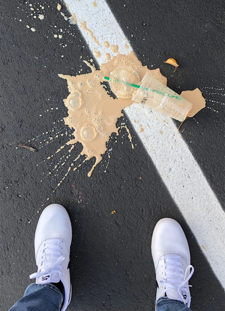 Een overgelopen plastic drinkbakje met een plastic bekertje met een straw en een bruine vloeistof, op een asfaltweg, met witte schoenen en blauwe jeans van de persoon die de foto heeft gemaakt.