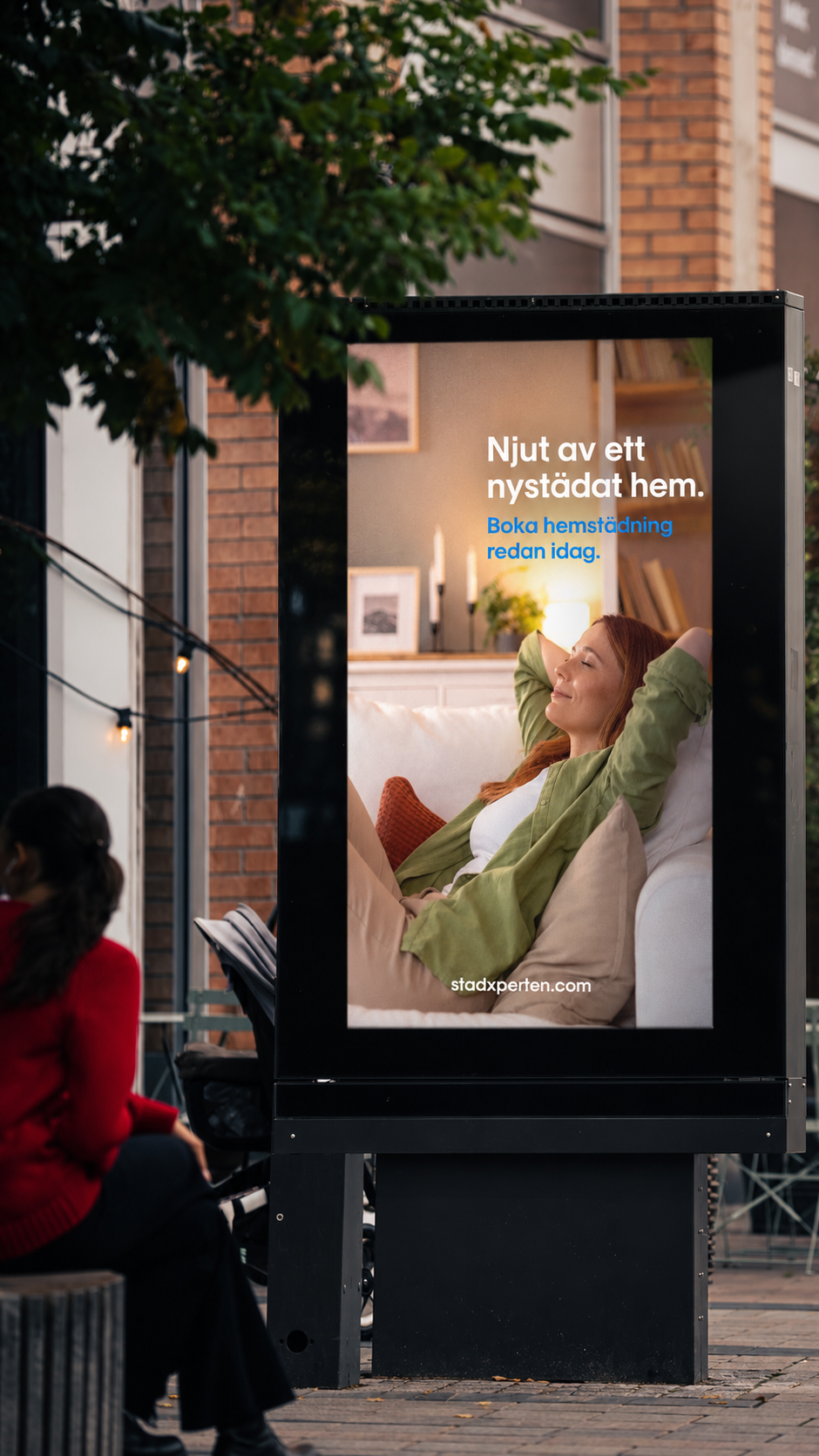 En digital annons på en utomhusskärm som visar en avslappnad kvinna som myser hemma, med texten 'Njut av ett nystädat hem. Boka hemstädning redan idag' och webbplatsen stadxperten.com.