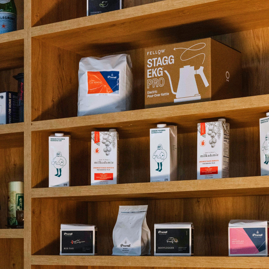 Wooden shelf filled with various boxes and packages, including boxes of Milkadamia unsweetened macadamia milk, a FELLOW Stagg EKG Pro electric kettle box, and other coffee-related products.