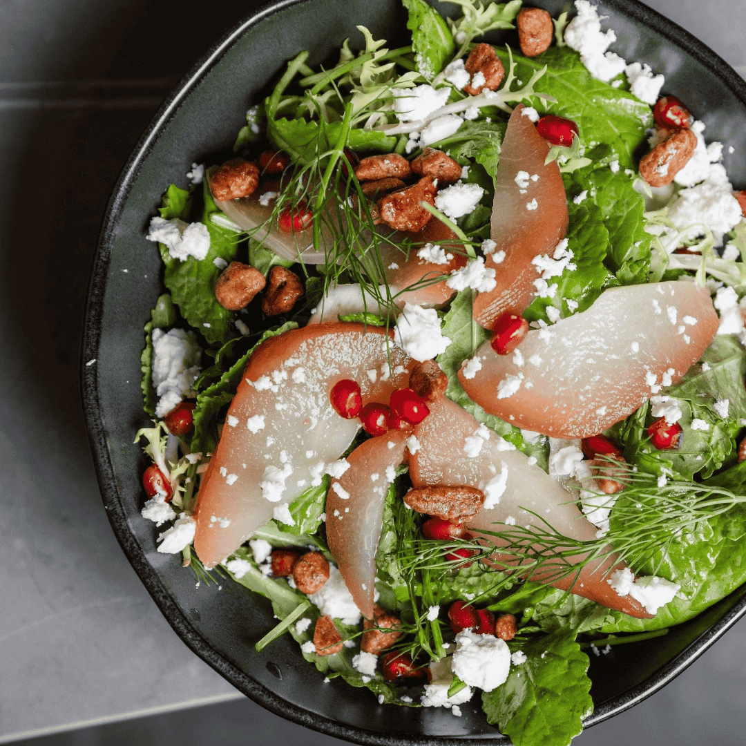 Fresh salad with mixed greens, sliced pears, pomegranate seeds, crumbled white cheese, toasted pecans, and fresh herbs in a black bowl.