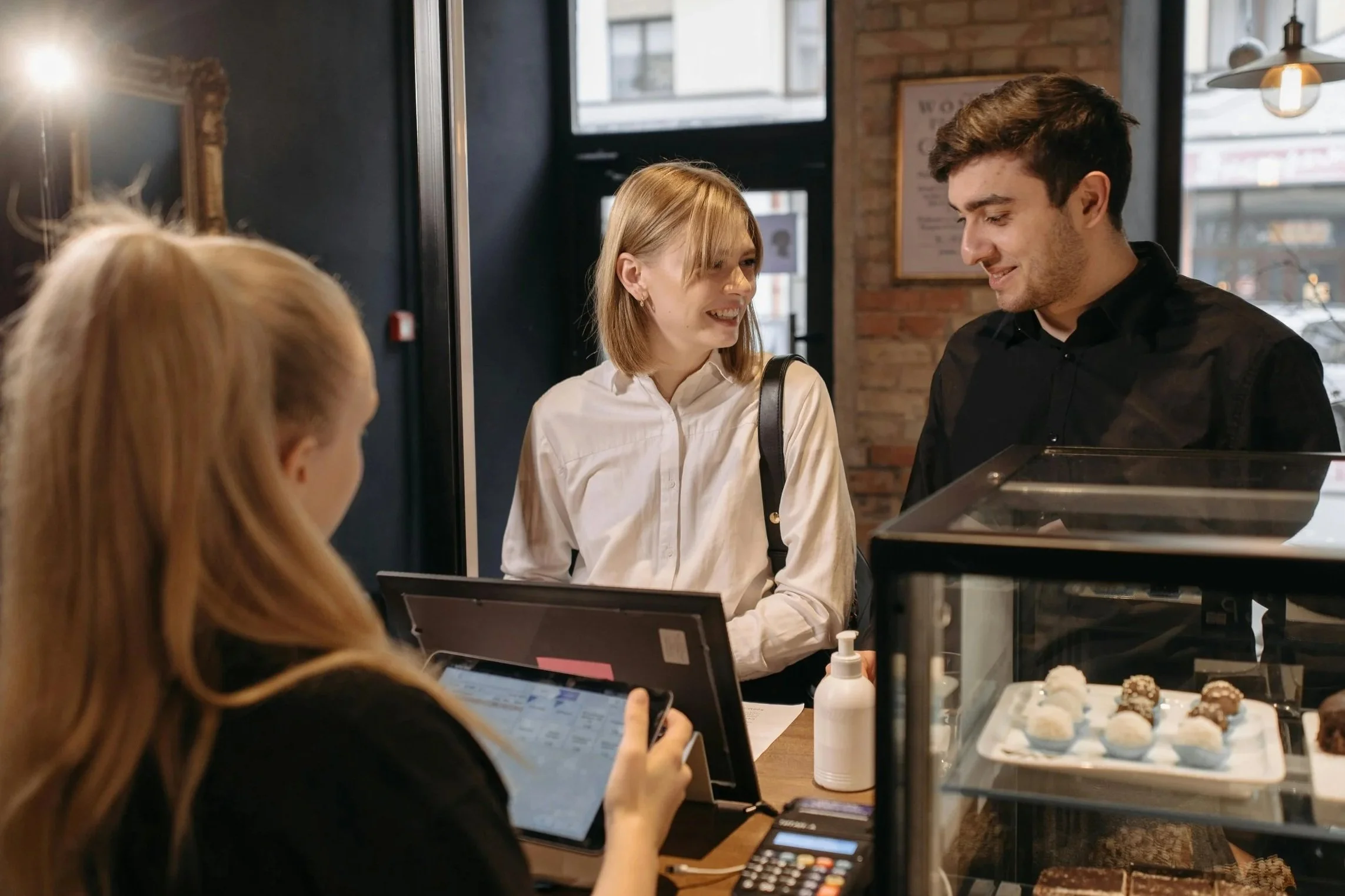 Couple ordering at a cafe counter with a barista using a tablet, next to a display case of desserts and a hand sanitizer bottle.