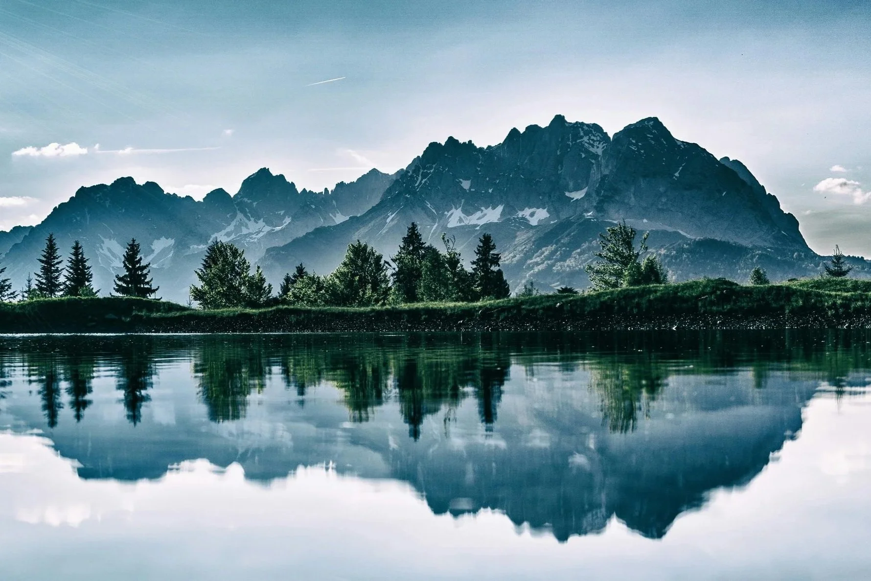 Mountainous landscape reflected in a calm lake, surrounded by pine trees under a bright sky, showcasing serene natural beauty.
