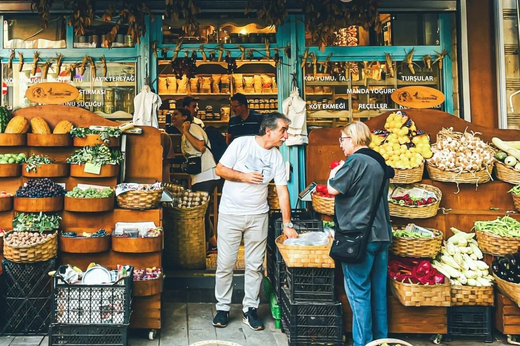 A bustling outdoor market with colorful produce and baskets, set against a vibrant orange wall and greenery.