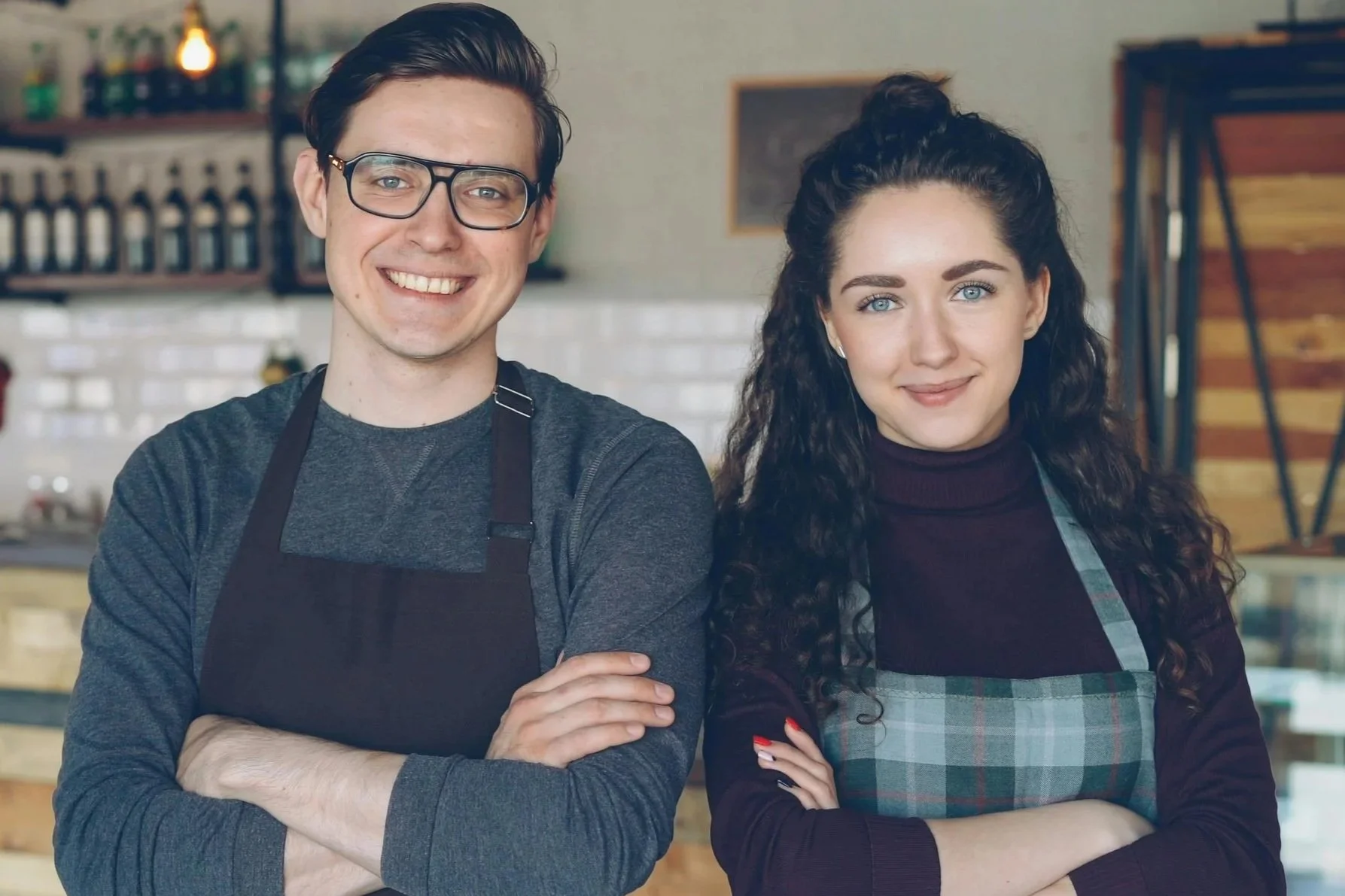 Two cafe workers standing side by side with arms crossed in front of a rustic wooden counter and shelves.