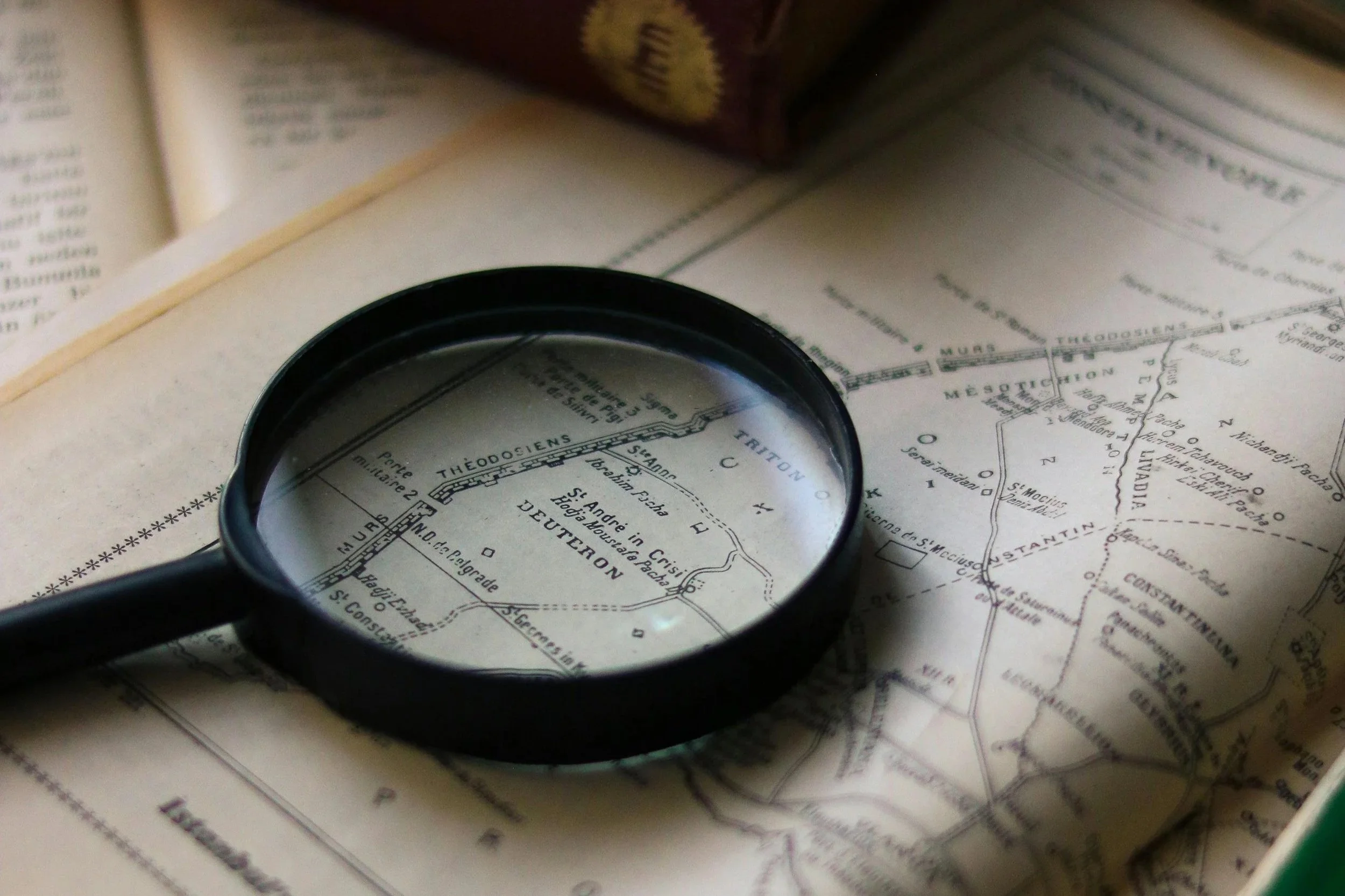 A magnifying glass rests on an open vintage map, surrounded by old books and pages, highlighting intricate geographic details.