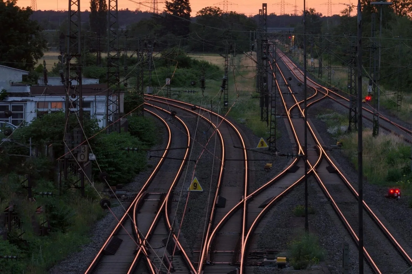 Railway tracks with glowing rails at sunset, surrounded by greenery and electrical poles under a vibrant orange sky.