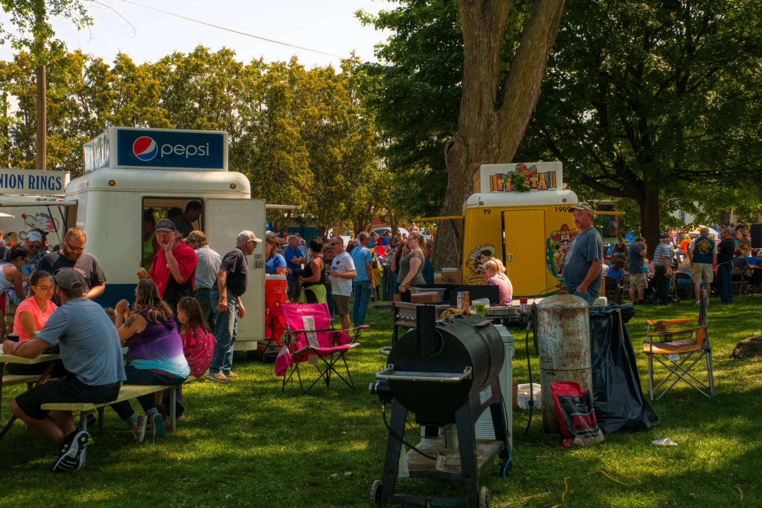 A bustling outdoor fair scene with food trucks, a crowd of people enjoying the event, and shaded picnic areas under trees.