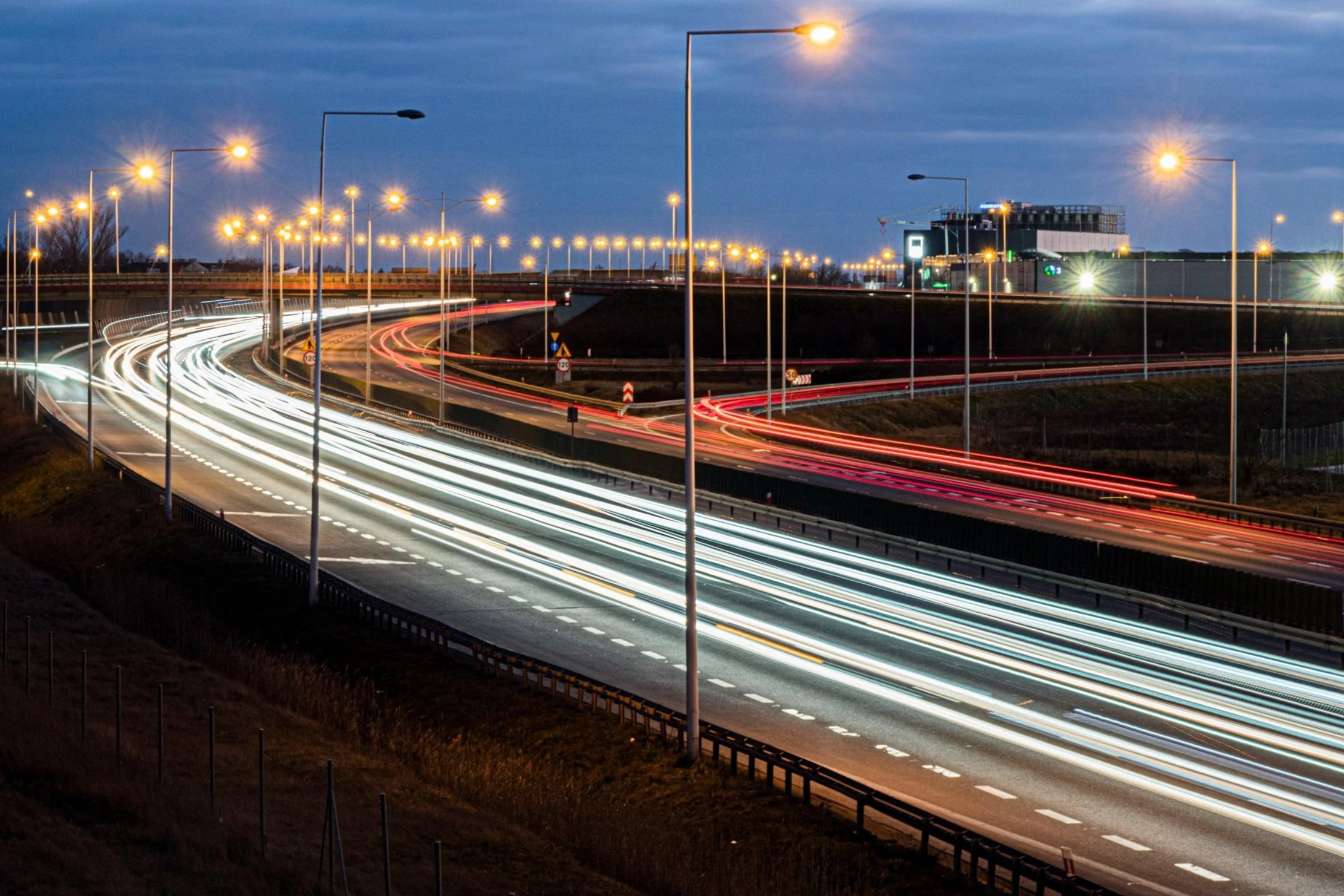 A long exposure shot of a highway at night, showcasing streaks of vehicle lights and illuminated street lamps against a cloudy sky.