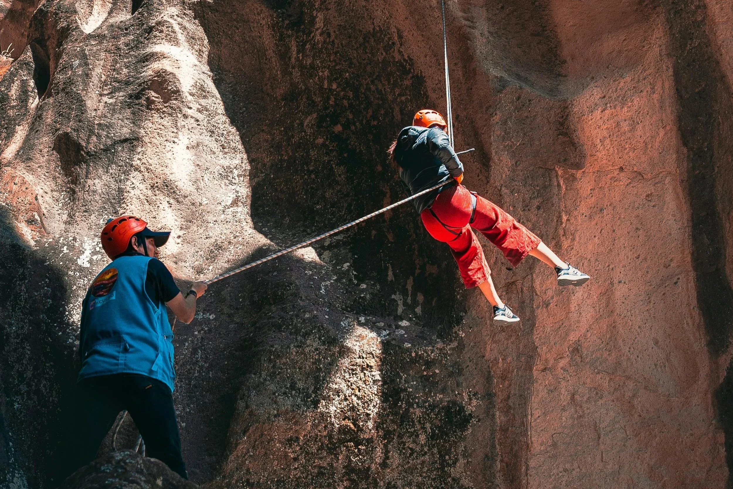 Two people rock climbing in a sunlit canyon, one ascending with ropes and the other belaying from below.