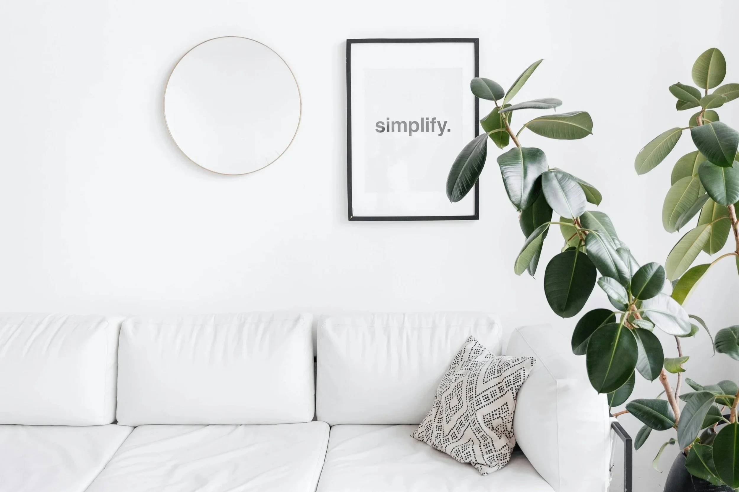 Minimalist white living room with a white sofa, patterned pillow, large green plant, round mirror, and framed "simplify." print on the wall.