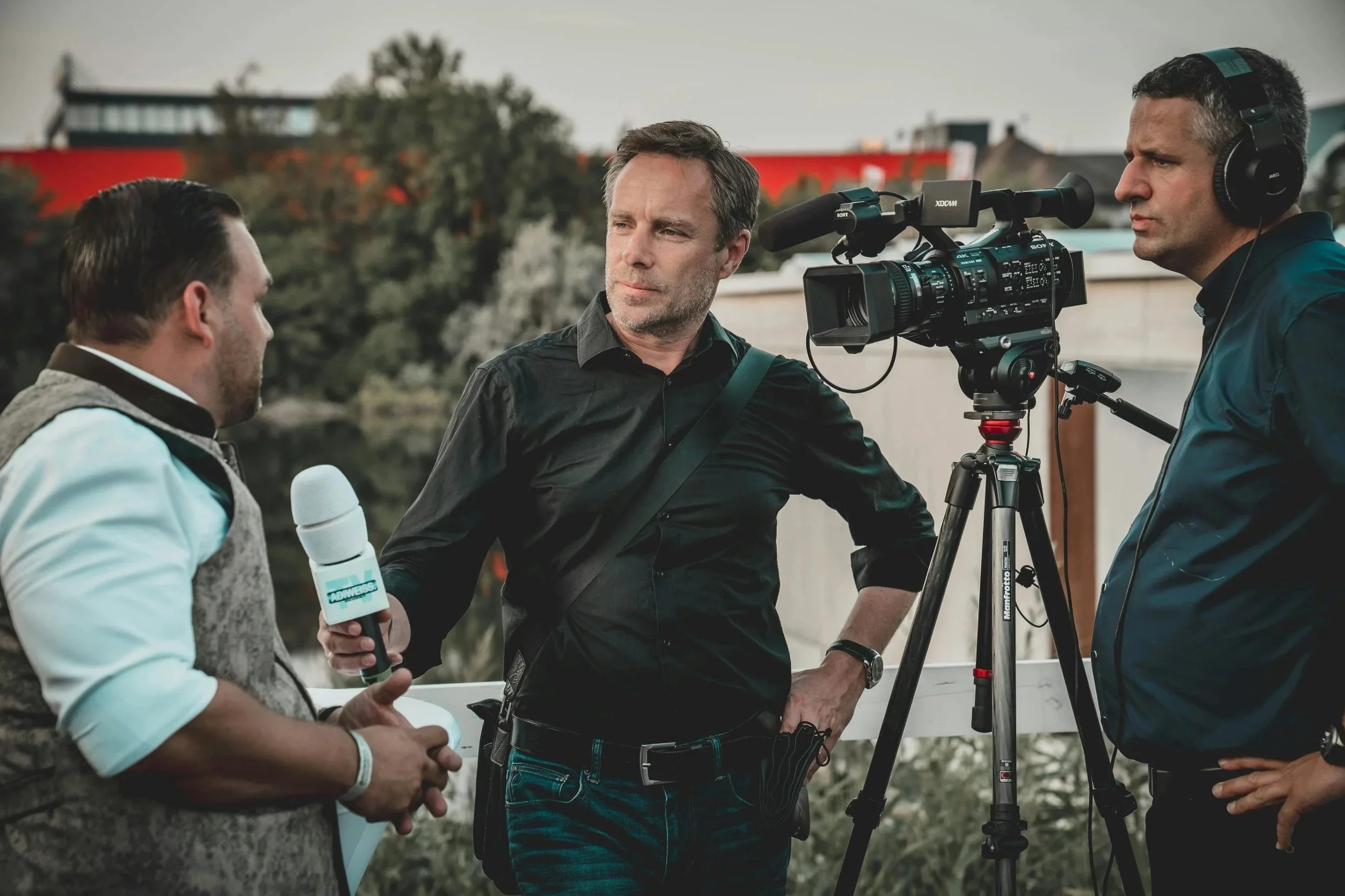 A camera crew interviews a man outdoors, with a camera and microphone setup, surrounded by greenery and a building in the background.