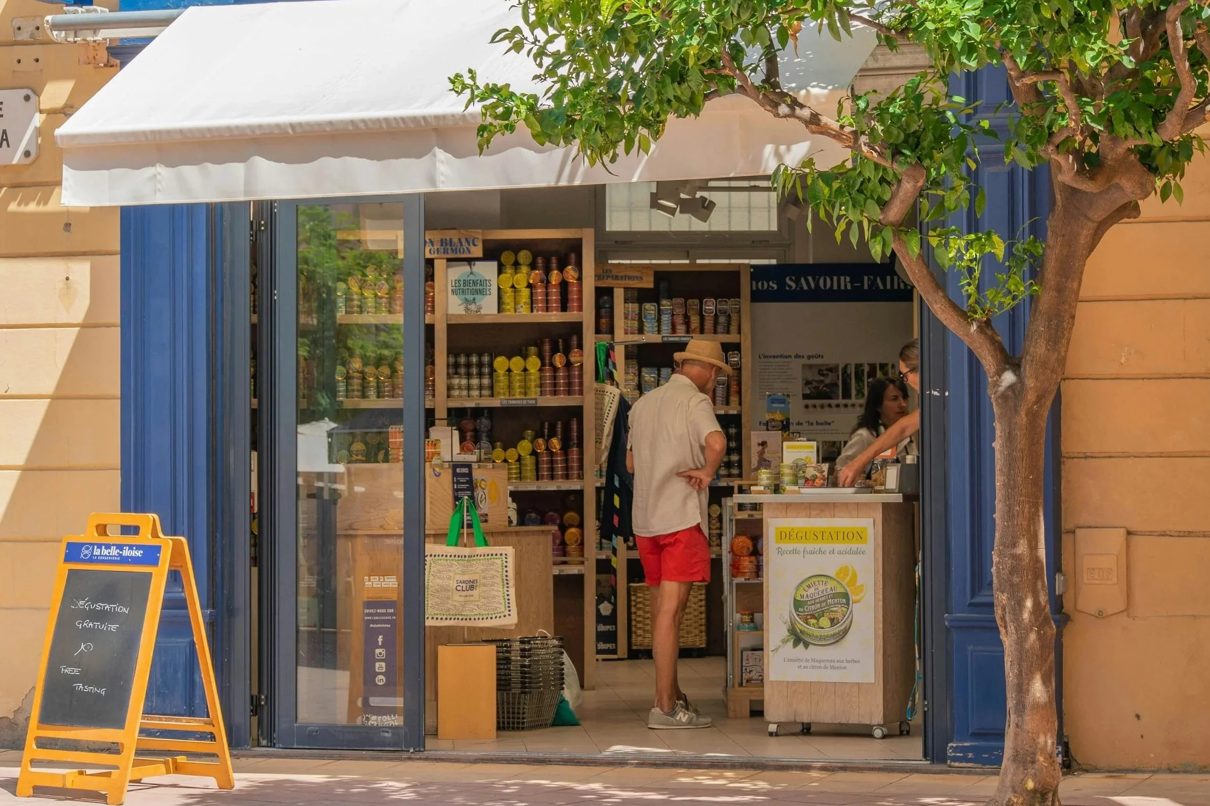 A sunny street scene featuring a shop named "La Conserverie" filled with colorful goods, a tree nearby, and a customer interacting inside.