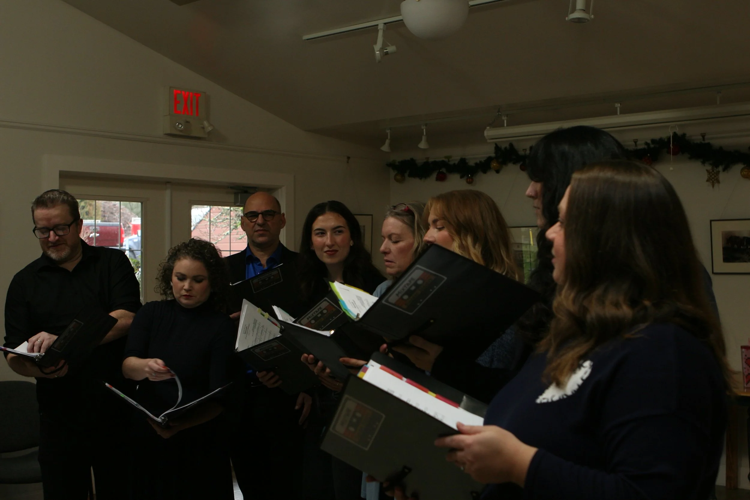Group of people singing or reading together indoors, holding sheet music or folders, with Christmas decorations on the wall.