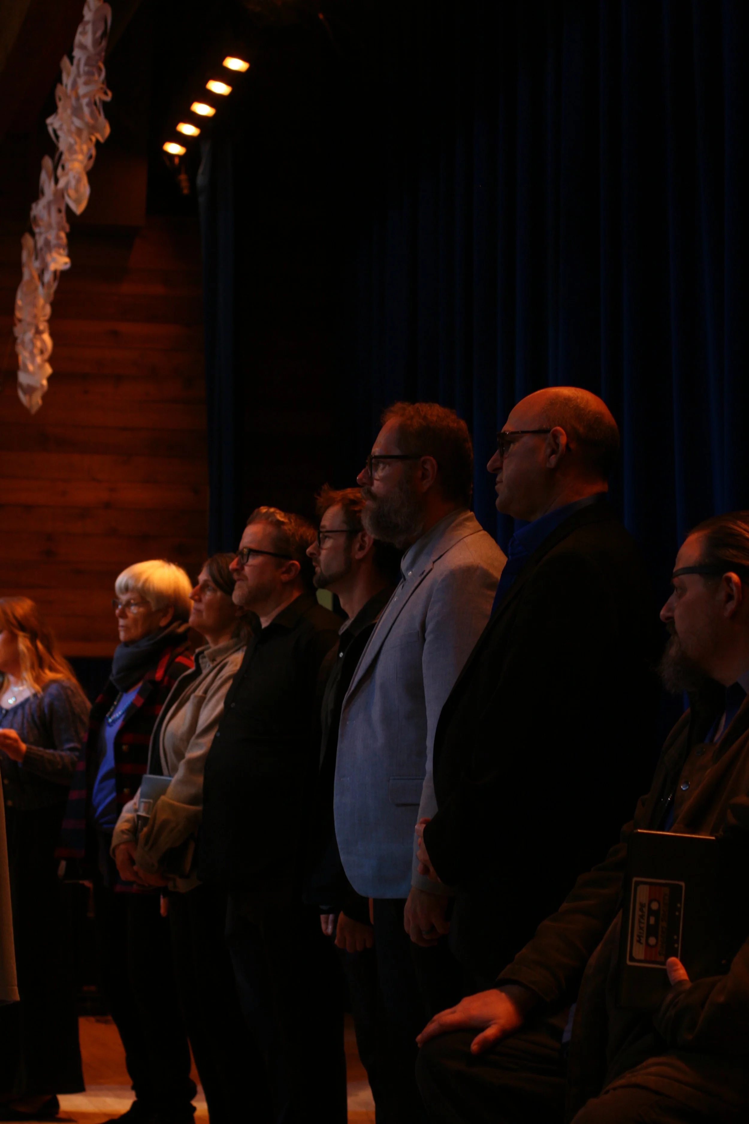 People standing in a row during an indoor event, with dark curtains behind them, some holding notebooks or papers, and a wooden wall and ceiling lights visible on the left.