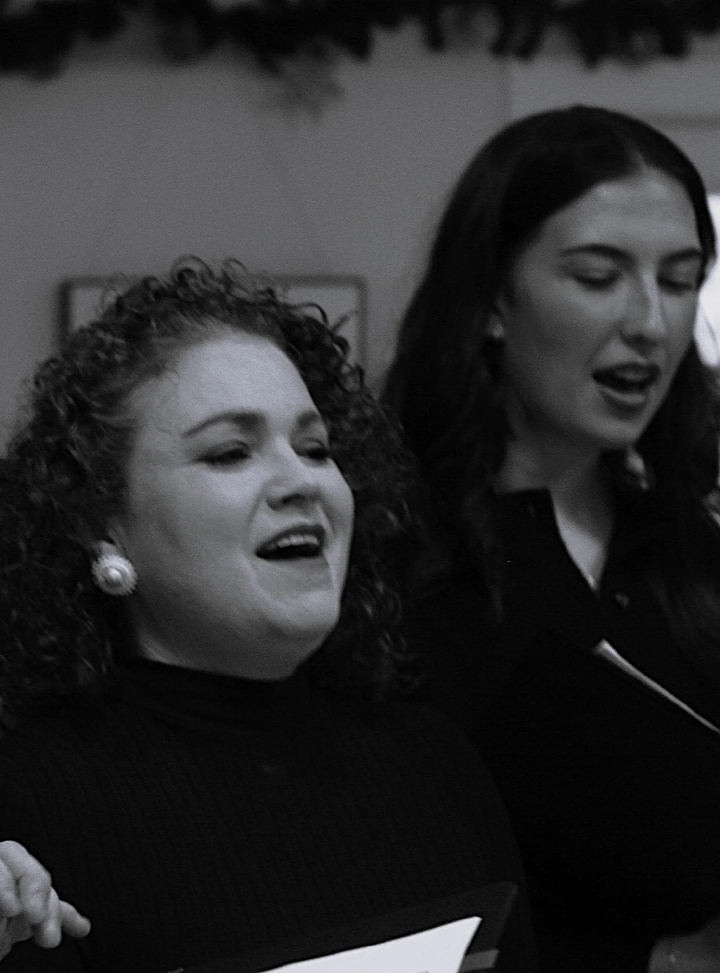 Two women with dark hair smiling and singing together, one with curly hair and pearl earrings, and the other with straight hair and earrings, in an indoor setting.