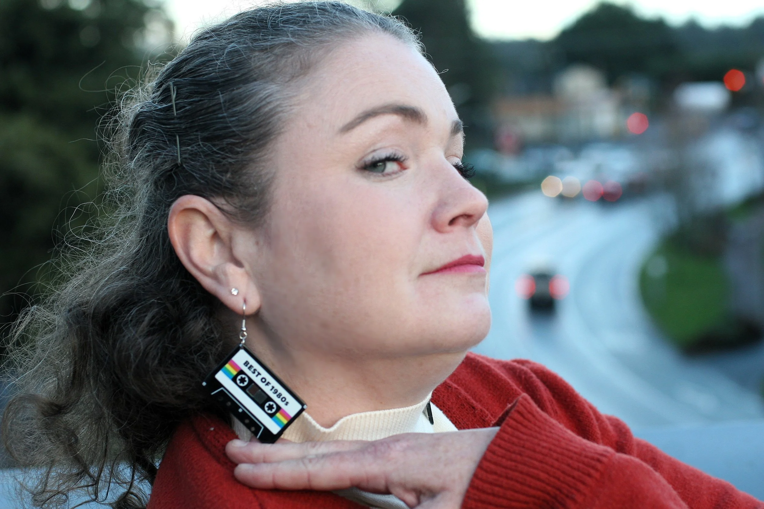 A woman with black hair, wearing a red sweater and a cream turtleneck, posing outdoors with a blurred road and cars in the background, wearing earrings including one shaped like a cassette tape with the text "Best of 1980s!".