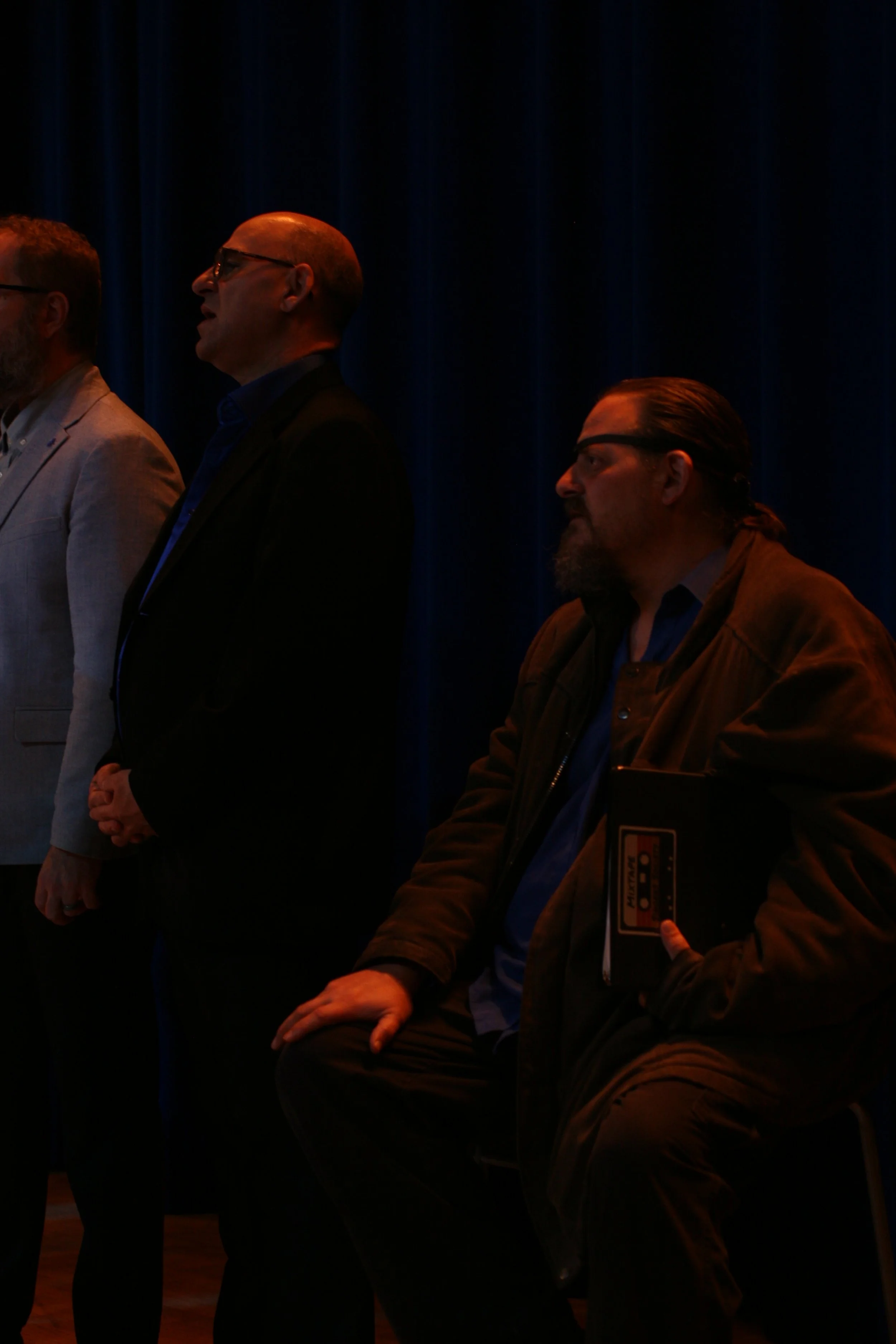 Three men standing and one man sitting on a chair in front of dark blue curtains.
