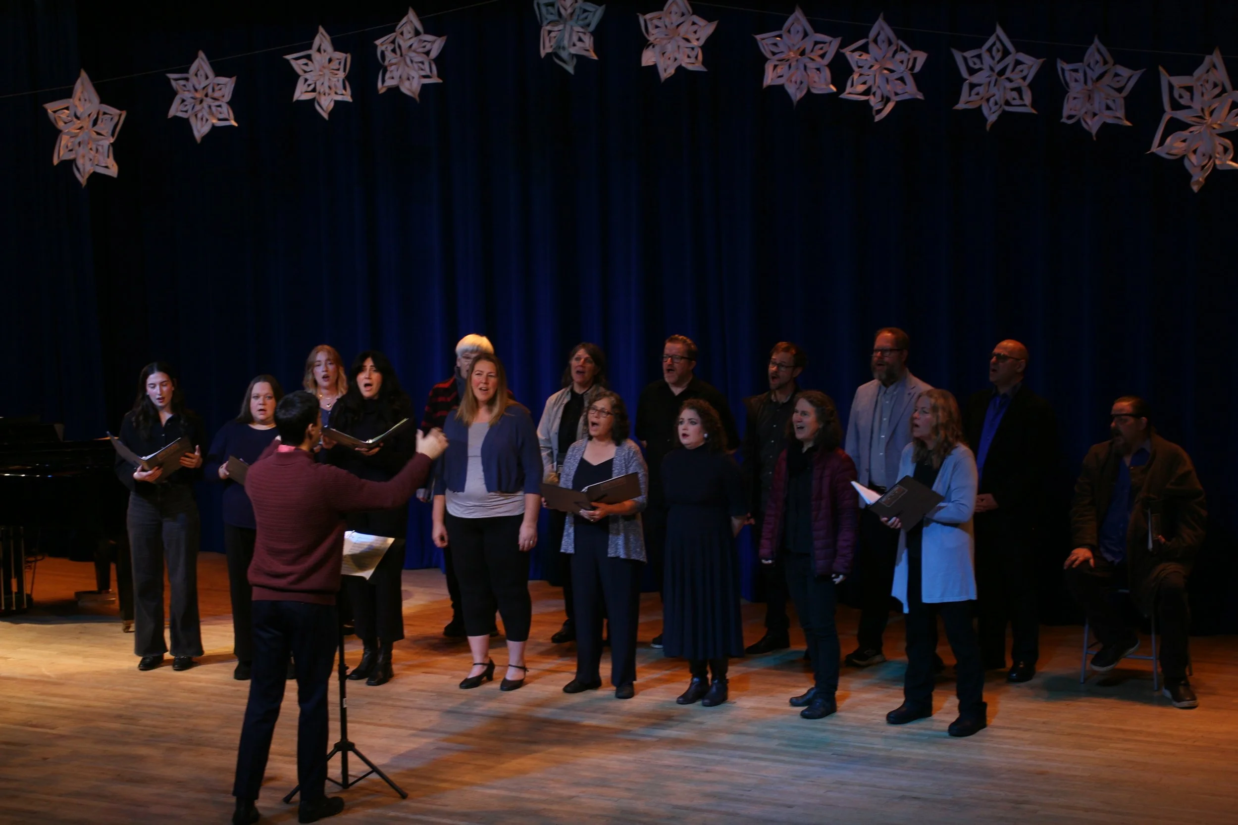 A choir group performing on stage with a conductor in front. The stage has dark blue curtains and hanging star-shaped decorations. The group includes men and women holding sheet music.