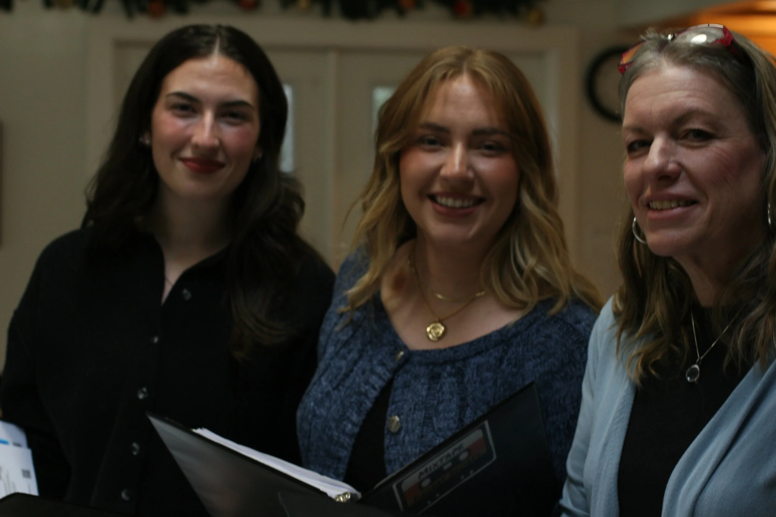Three women smiling in an indoor setting, holding documents or folders.
