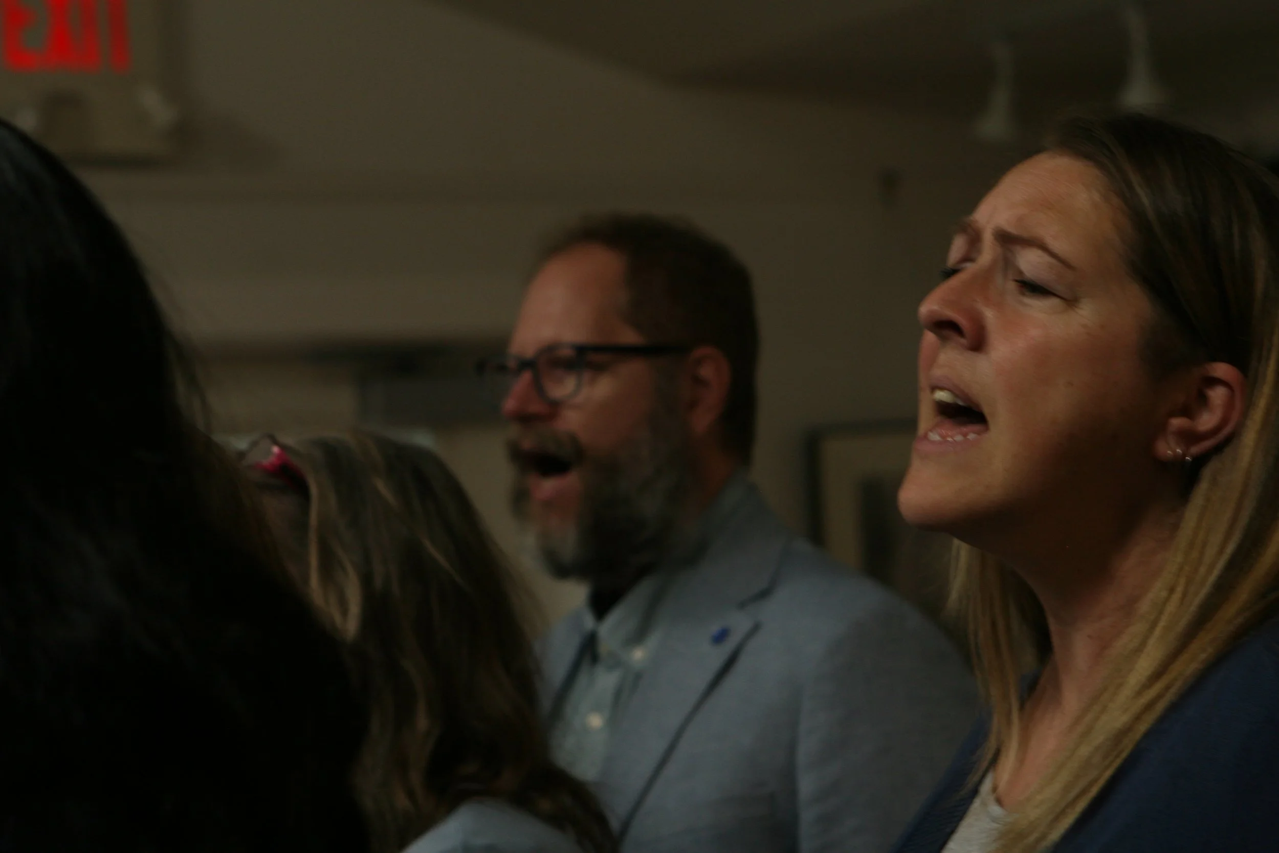 People singing or speaking, with one woman in the foreground and a man in the background, in a close-up indoor setting.