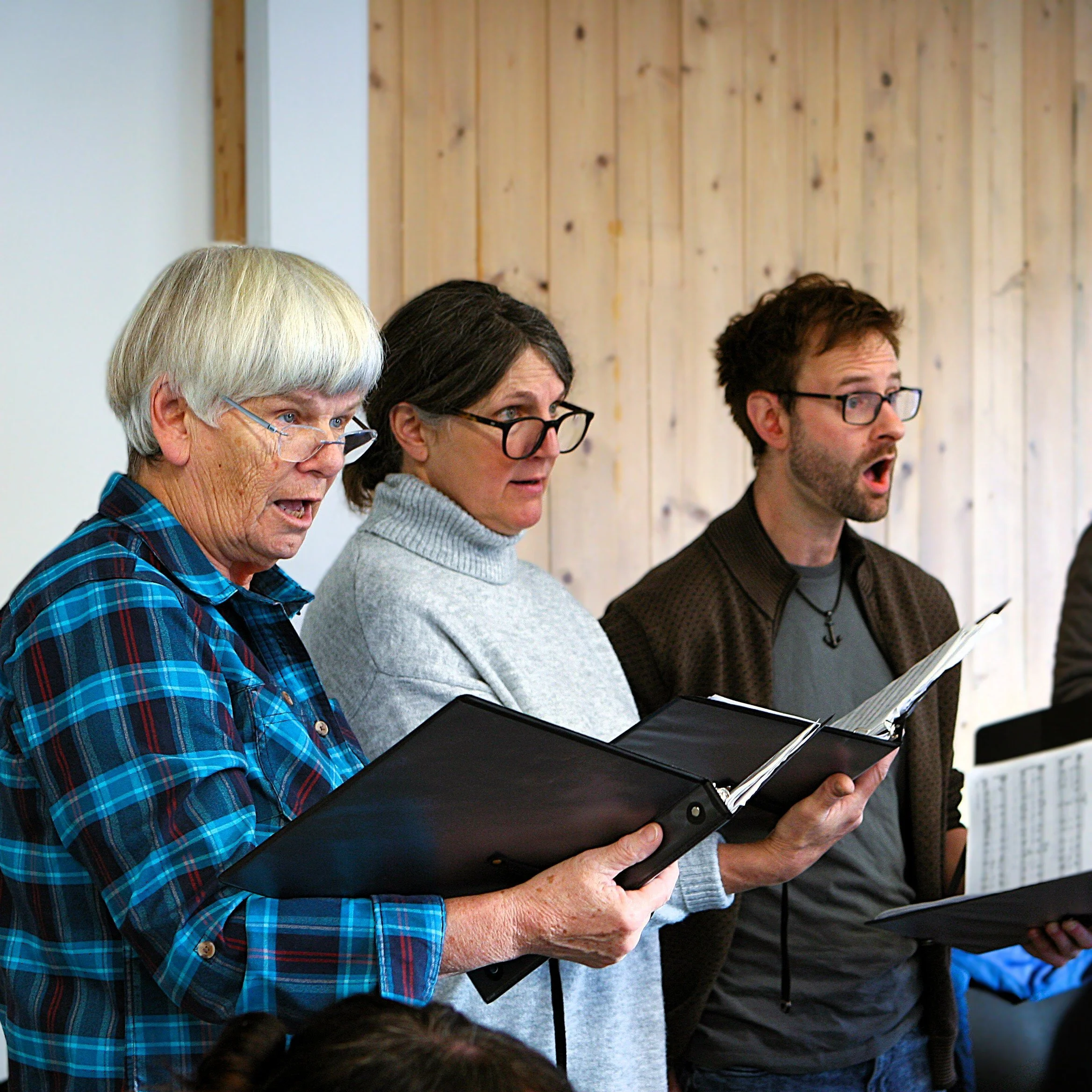 Three people singing or rehearsing with songbooks in hand, standing in a room with wooden wall paneling.