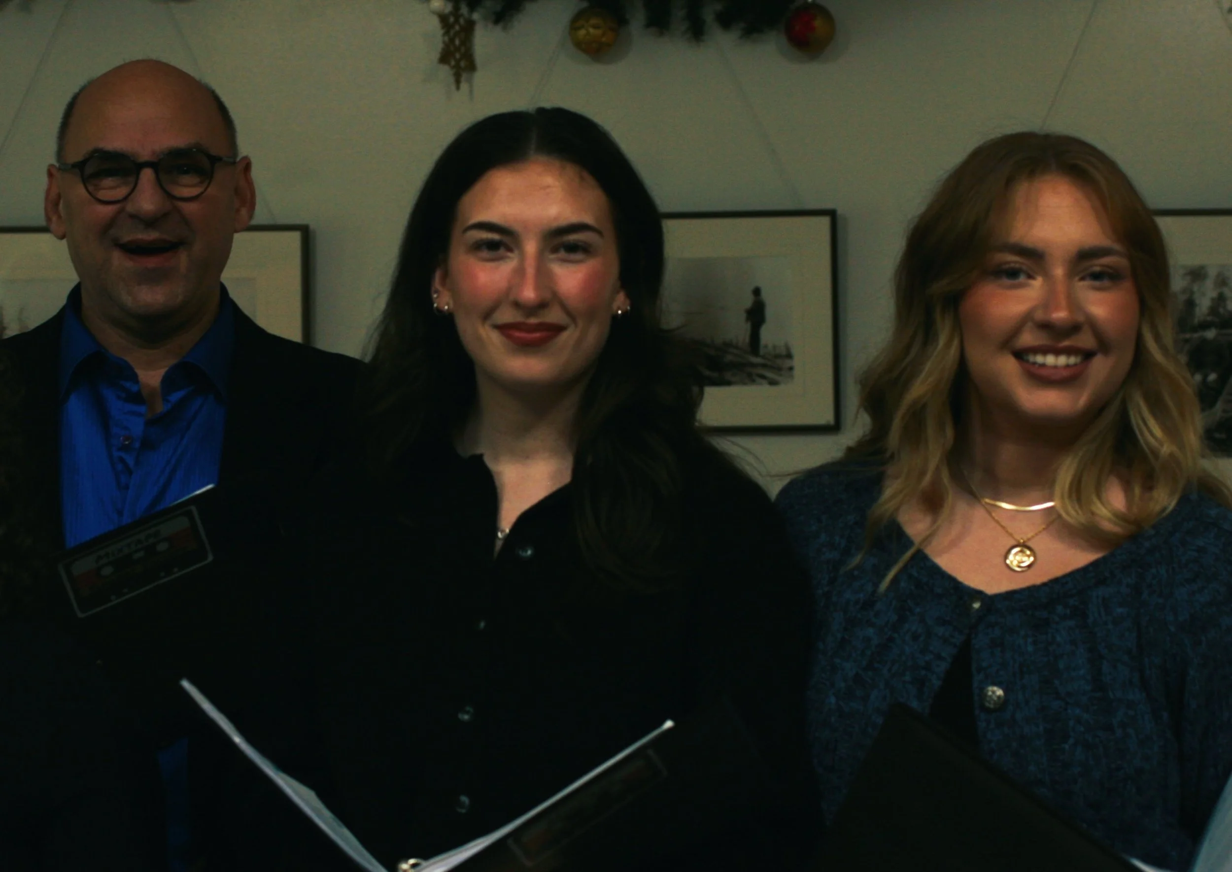 Three people standing close together indoors, two women in the center and right, and a man on the left. They are smiling and dressed in semi-formal attire. The background has framed pictures on the wall and some decorations hanging from the ceiling.