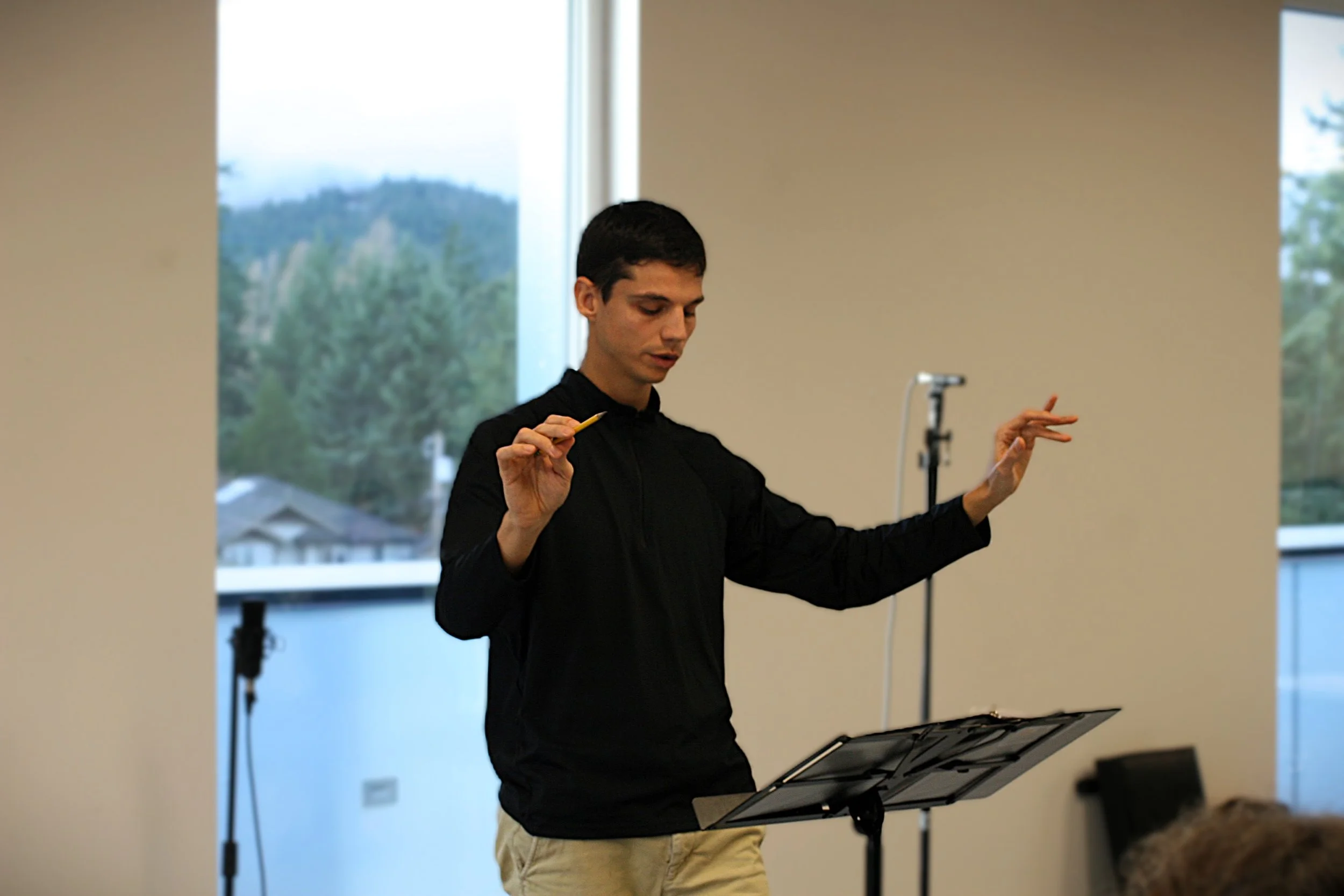 A young man conducting or leading a musical performance indoors with a music stand in front of him, large window in the background showing trees and a cloudy sky.