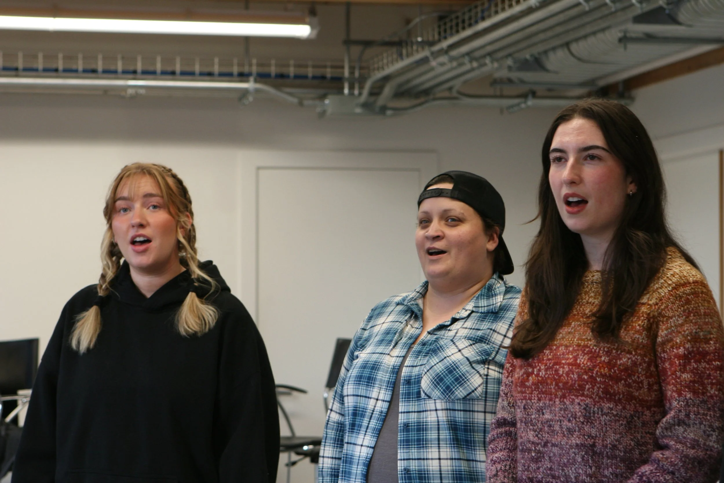 Three women singing together in a room with computers and exposed pipes in the ceiling.