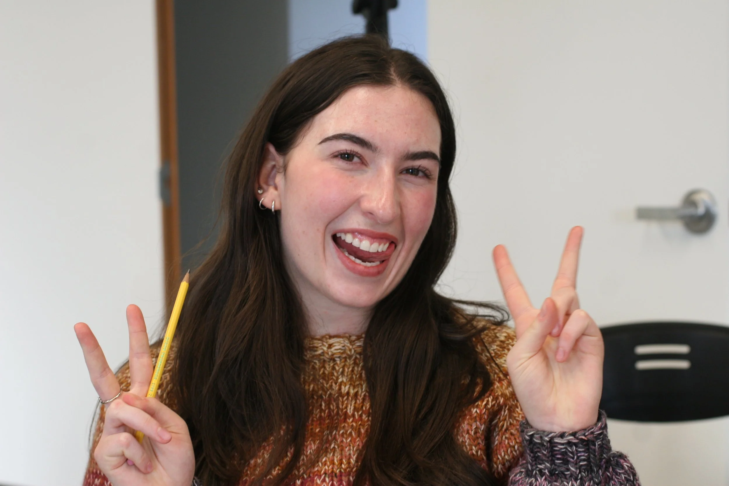 A young woman with long dark hair and earrings smiling and making peace signs with both hands while holding a pencil.