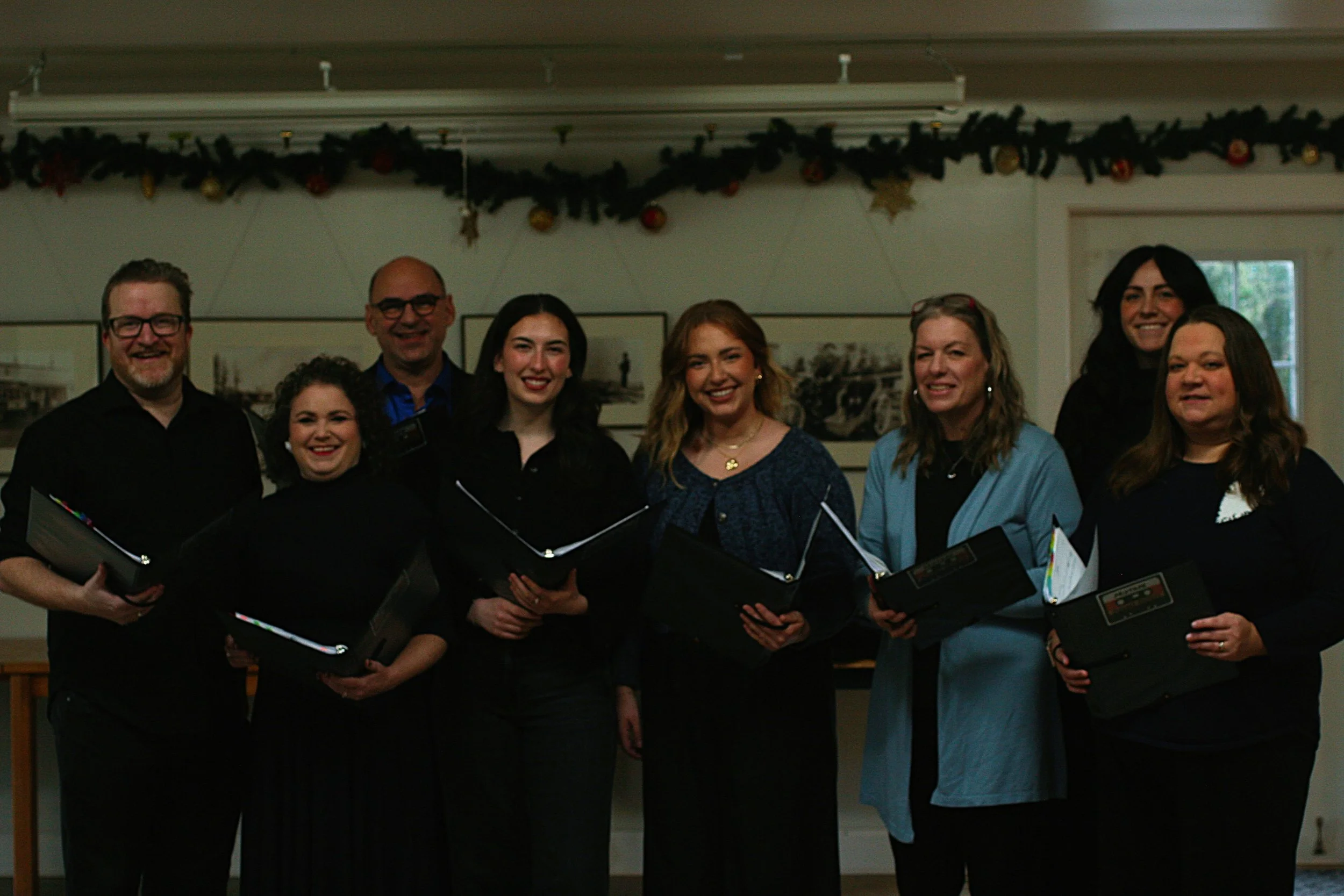 Group of eight people, five women and three men, standing together indoors, smiling, holding black folders, in front of a wall decorated with holiday garland and artwork, with a window on the right.