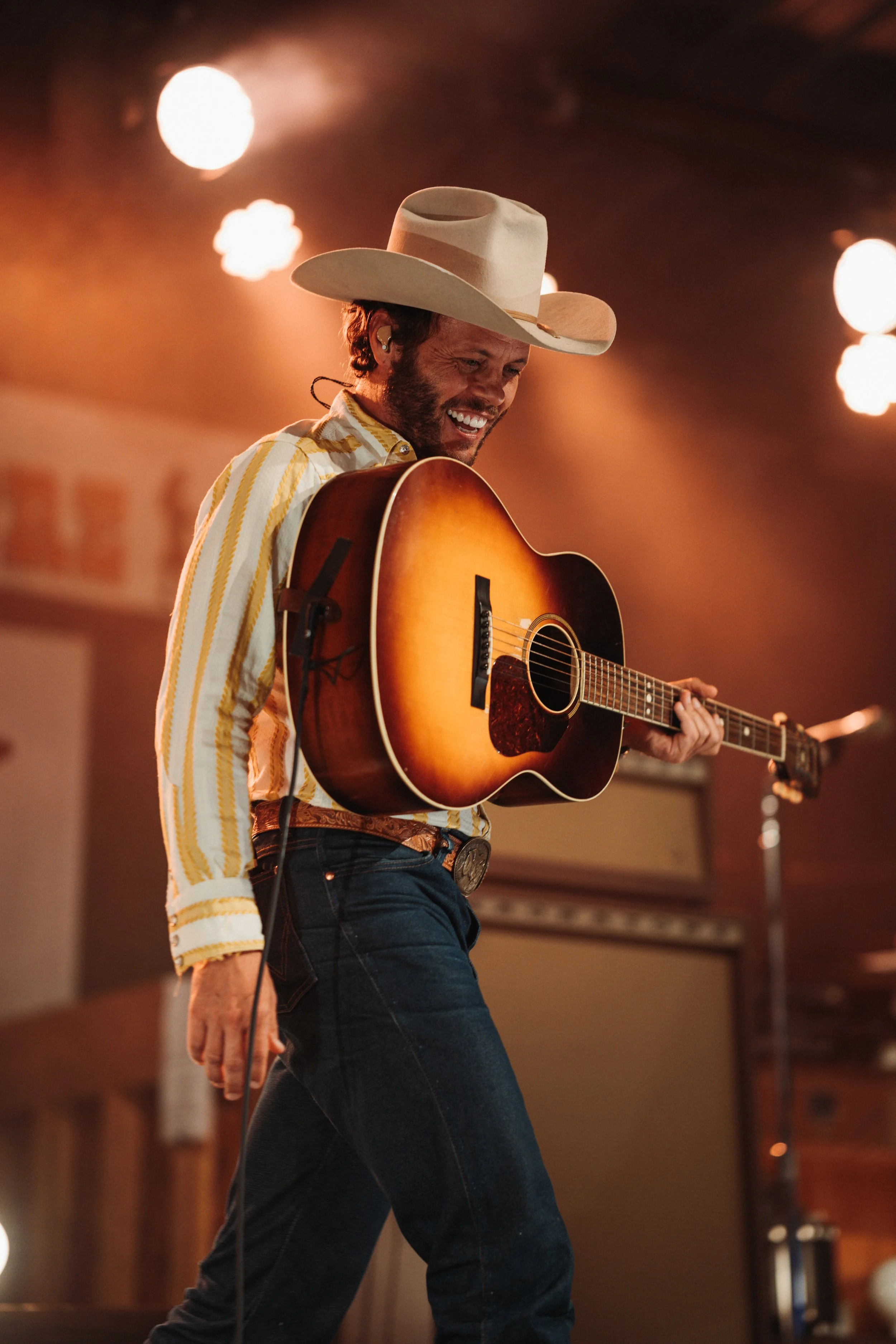 Charley Crockett | Man wearing a cowboy hat and striped shirt, playing an acoustic guitar on stage, smiling.
