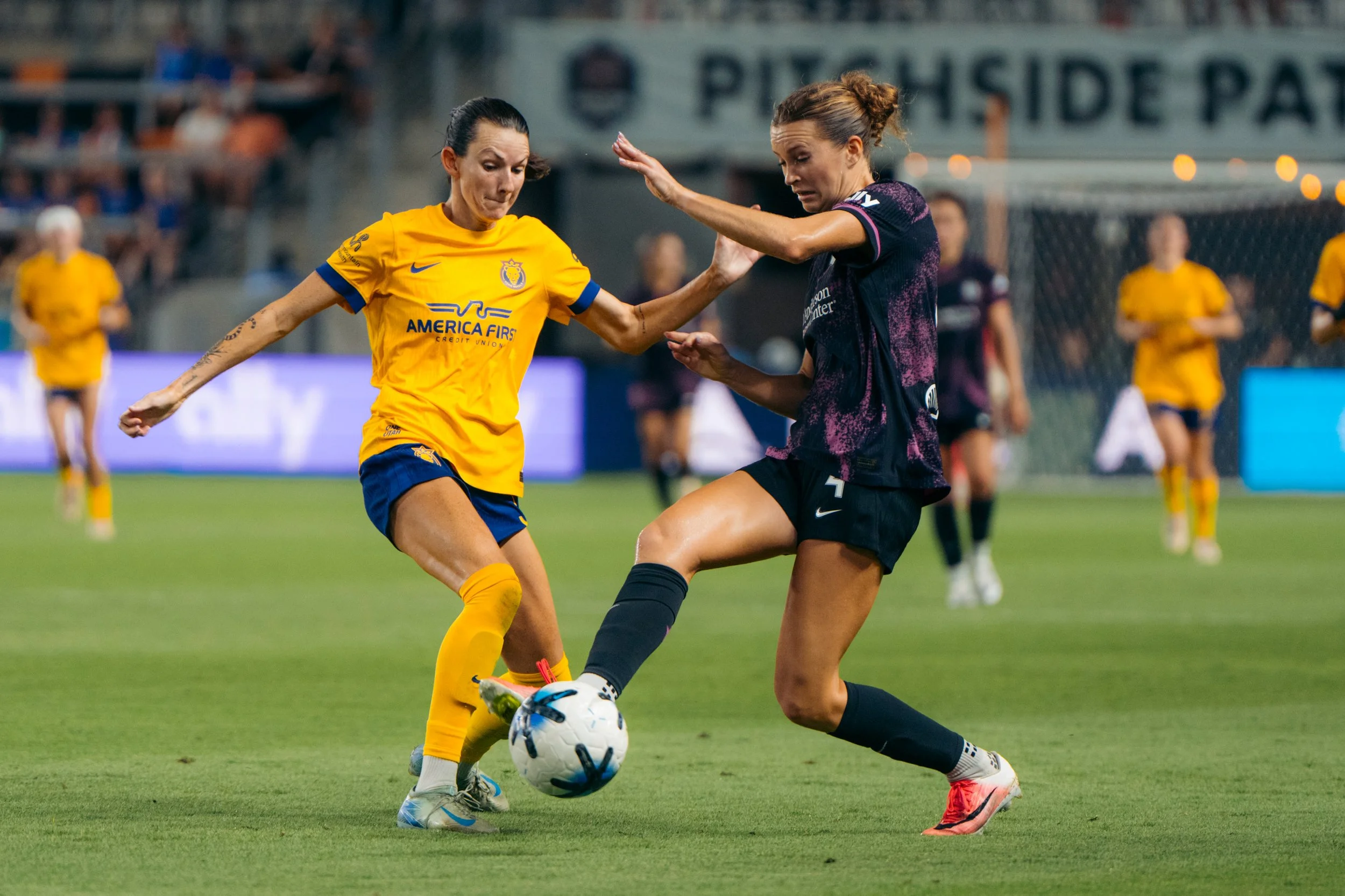 Houston Dash | Two female soccer players competing for ball on field, one in yellow and the other in dark jersey, with blurred spectators in background.