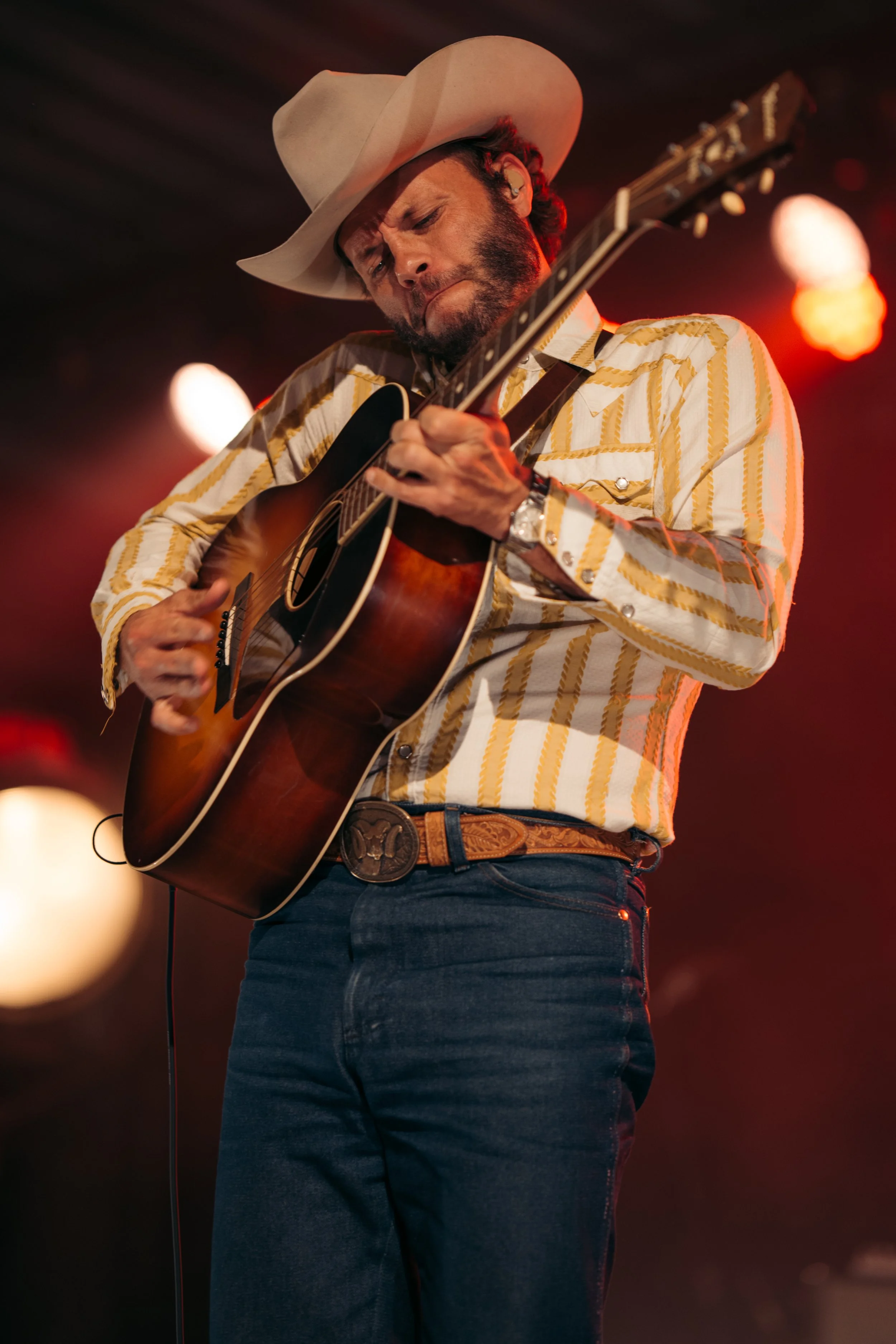 Charley Crockett | A man in a cowboy hat, striped shirt, and jeans playing an acoustic guitar on stage with clips of red and orange lights in the background.