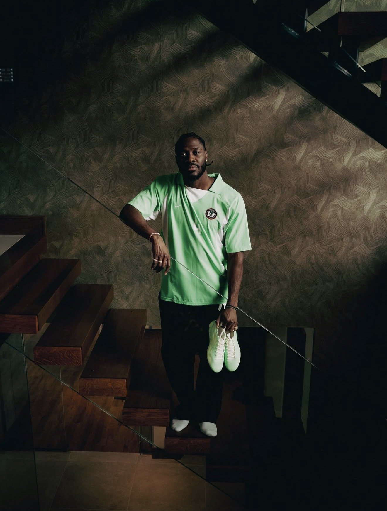A man stands on a staircase holding a pair of white athletic shoes in his left hand and a fishing rod in his right. He is wearing a green soccer jersey, black pants, and white socks. The background features a textured wall with a shadowed, greenish ambiance.